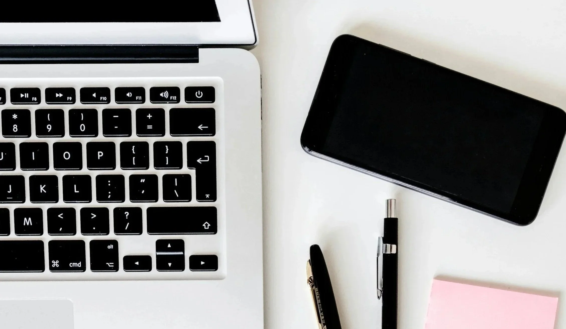 A white computer keyboard, a gold pen, a pink-purplish pen, round glasses, and an open notebook on a white desk.