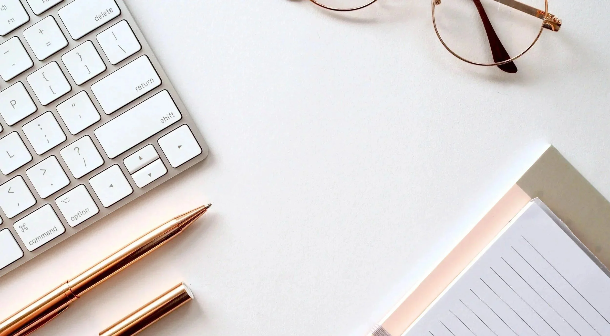 A white computer keyboard, a gold pen, a pink-purplish pen, round glasses, and an open notebook on a white desk.
