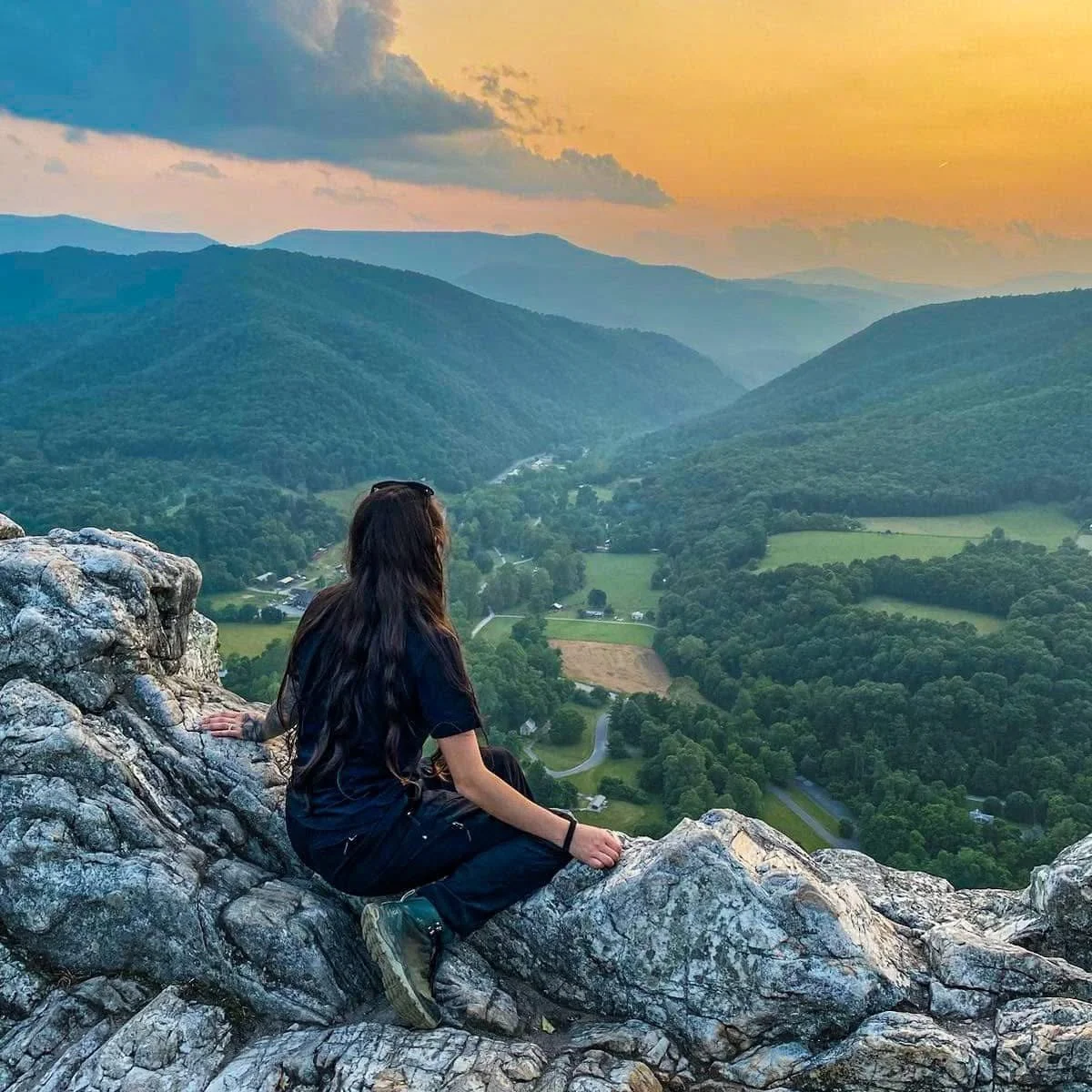 A woman sitting on rocky ledge overlooking a green valley with mountains at sunset.