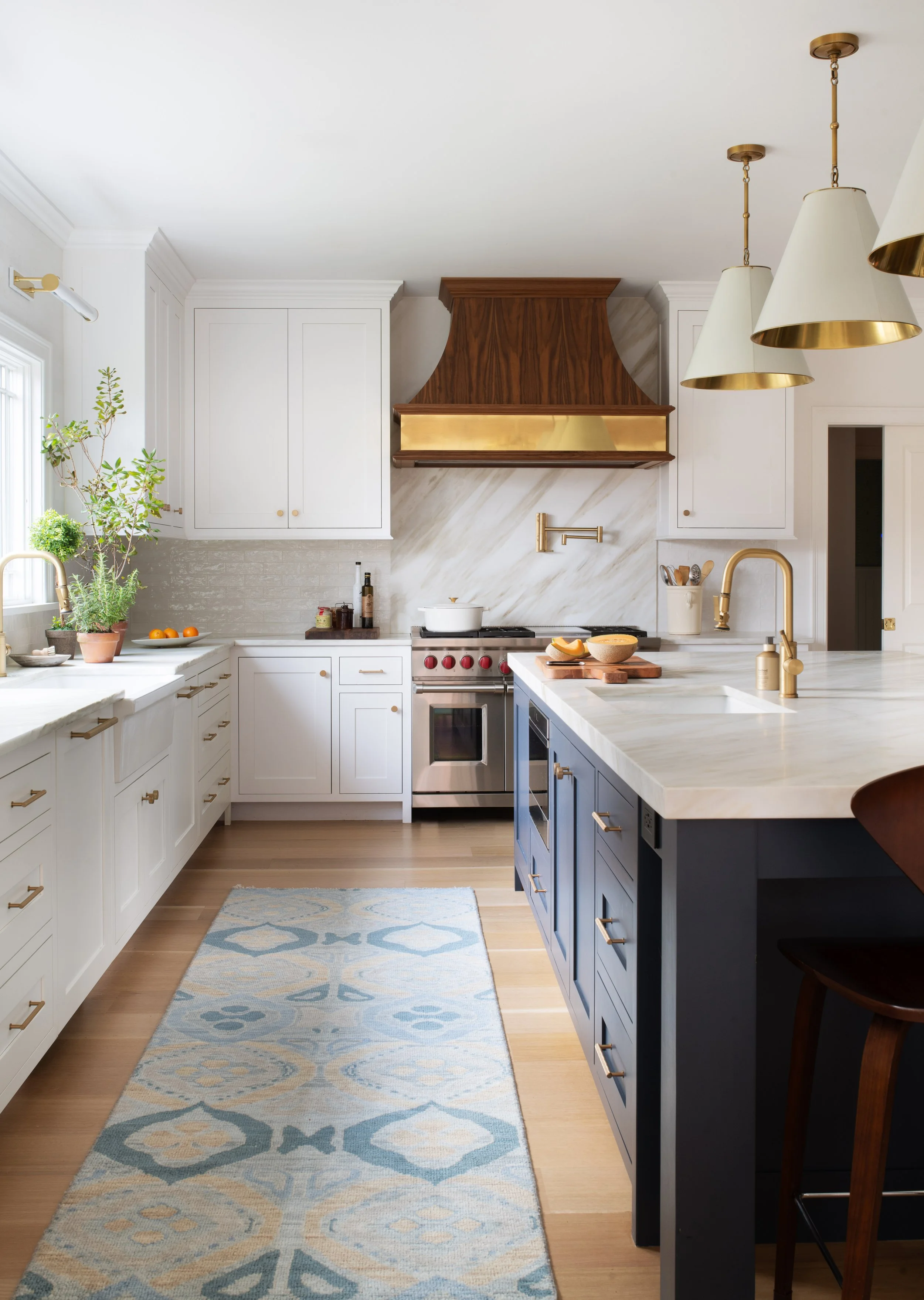 A modern kitchen with white and navy cabinets, a kitchen island, gold fixtures, a white countertop, open shelving, plants, and a window with natural light.