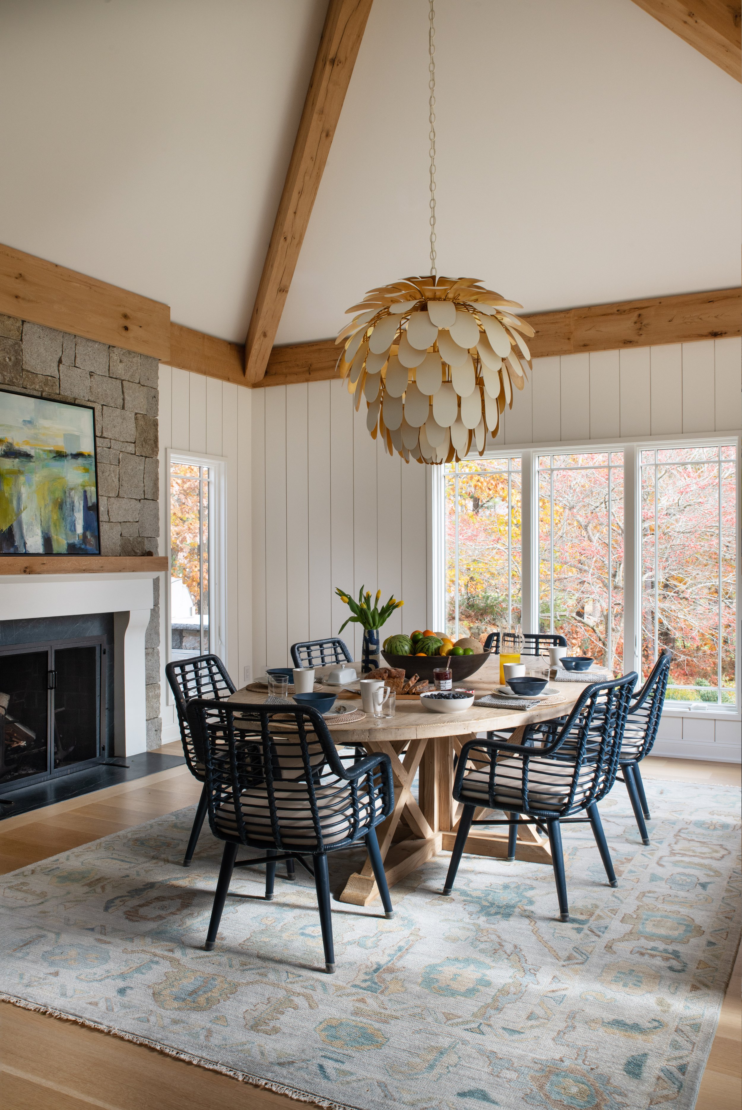 A cozy dining room with a large round wooden table, black chairs, and a colorful fruit bowl. The room features a stone fireplace, a large abstract painting, white shiplap walls, and large windows with a view of autumn trees. A unique chandelier hangs from the ceiling.