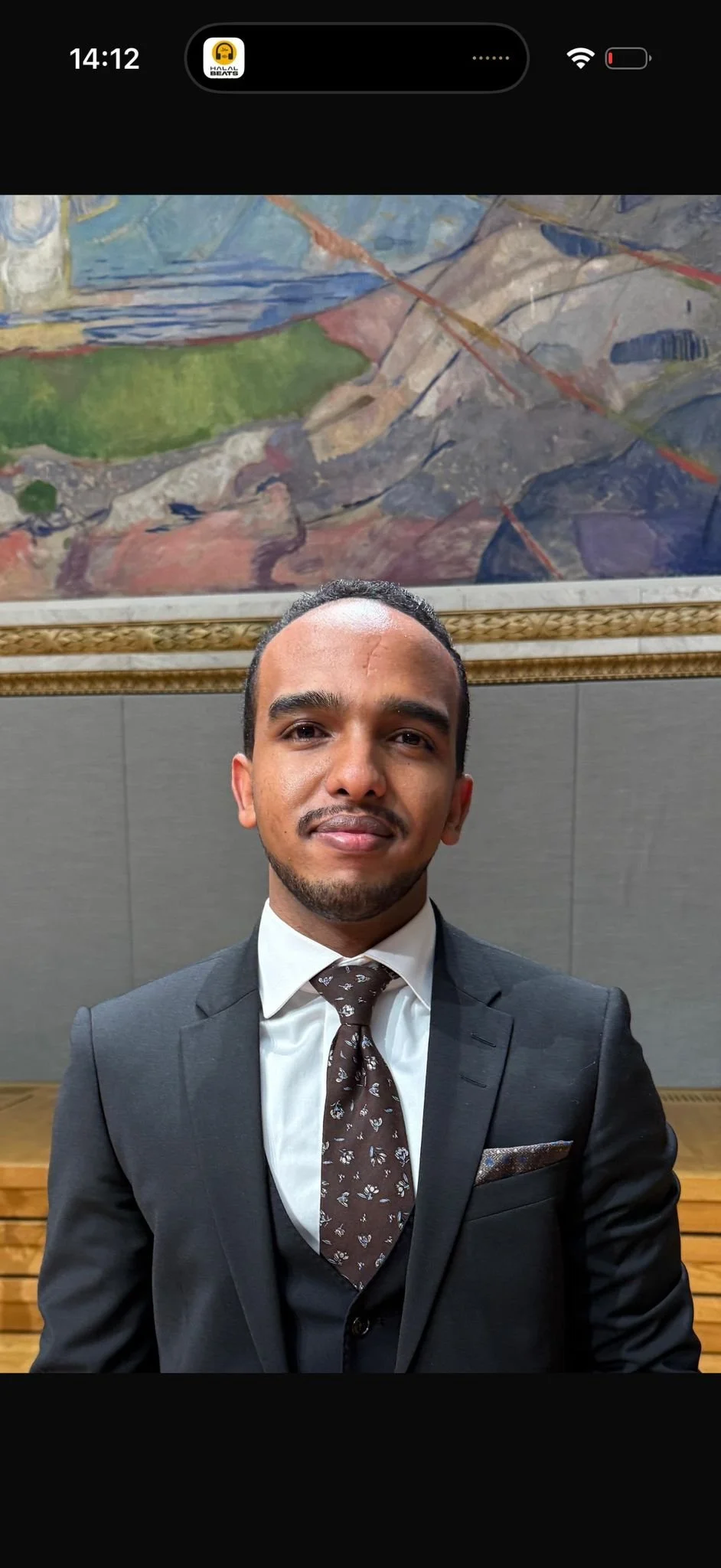 Nærbilde av en ung mann med mørk hud, kort krøllete hår og iført en svart og grå rutete skjorte, som står foran en lys vegg.