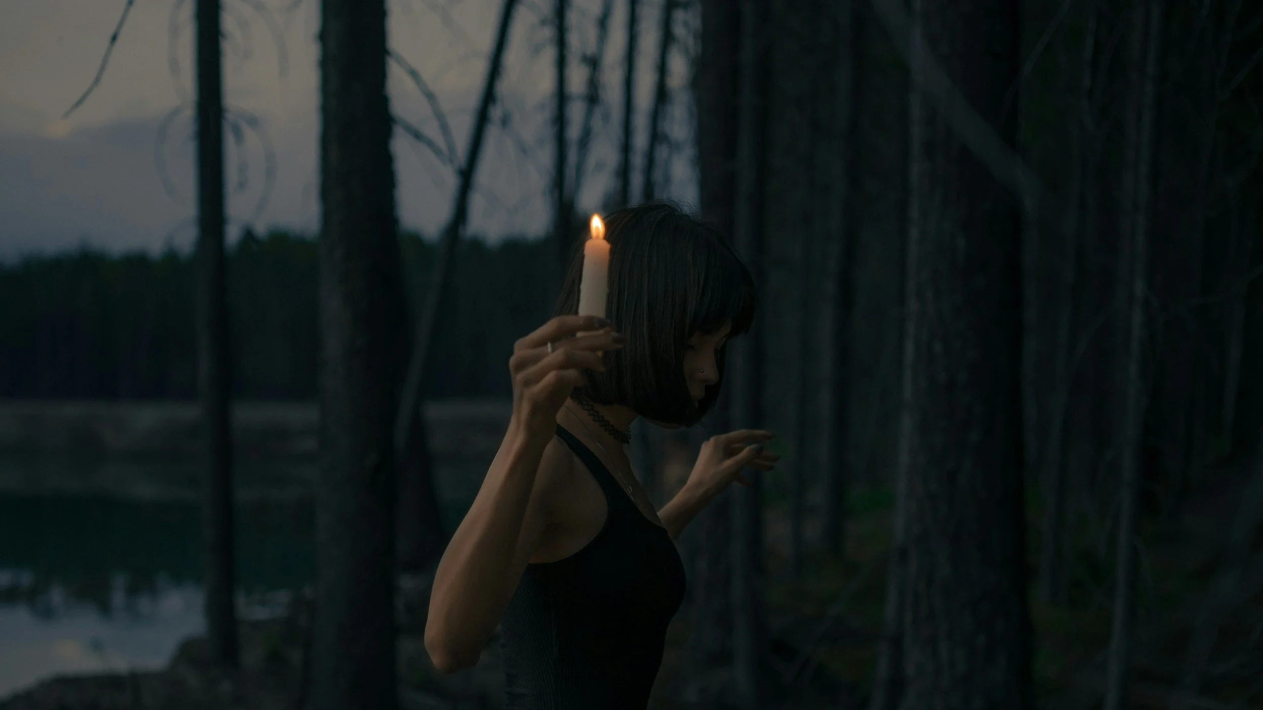 A woman with dark hair and a black tank top standing in a dark wooded area holding a lit candle near her head.