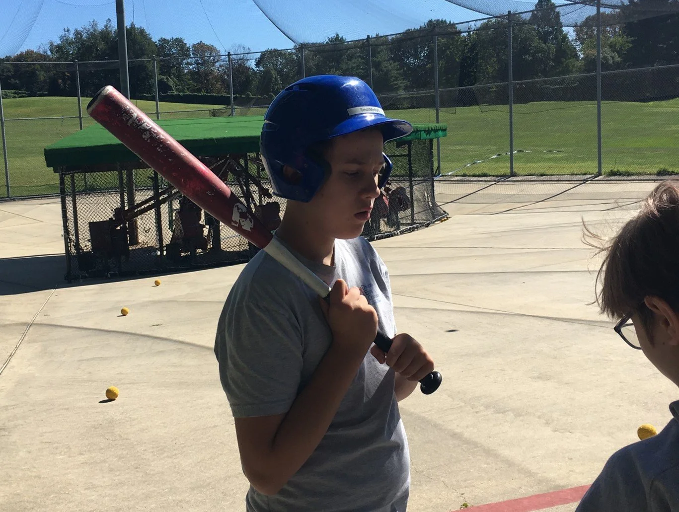 A young boy wearing a blue helmet holds a red baseball bat on a baseball field, with a green and black dugout and baseball equipment in the background.