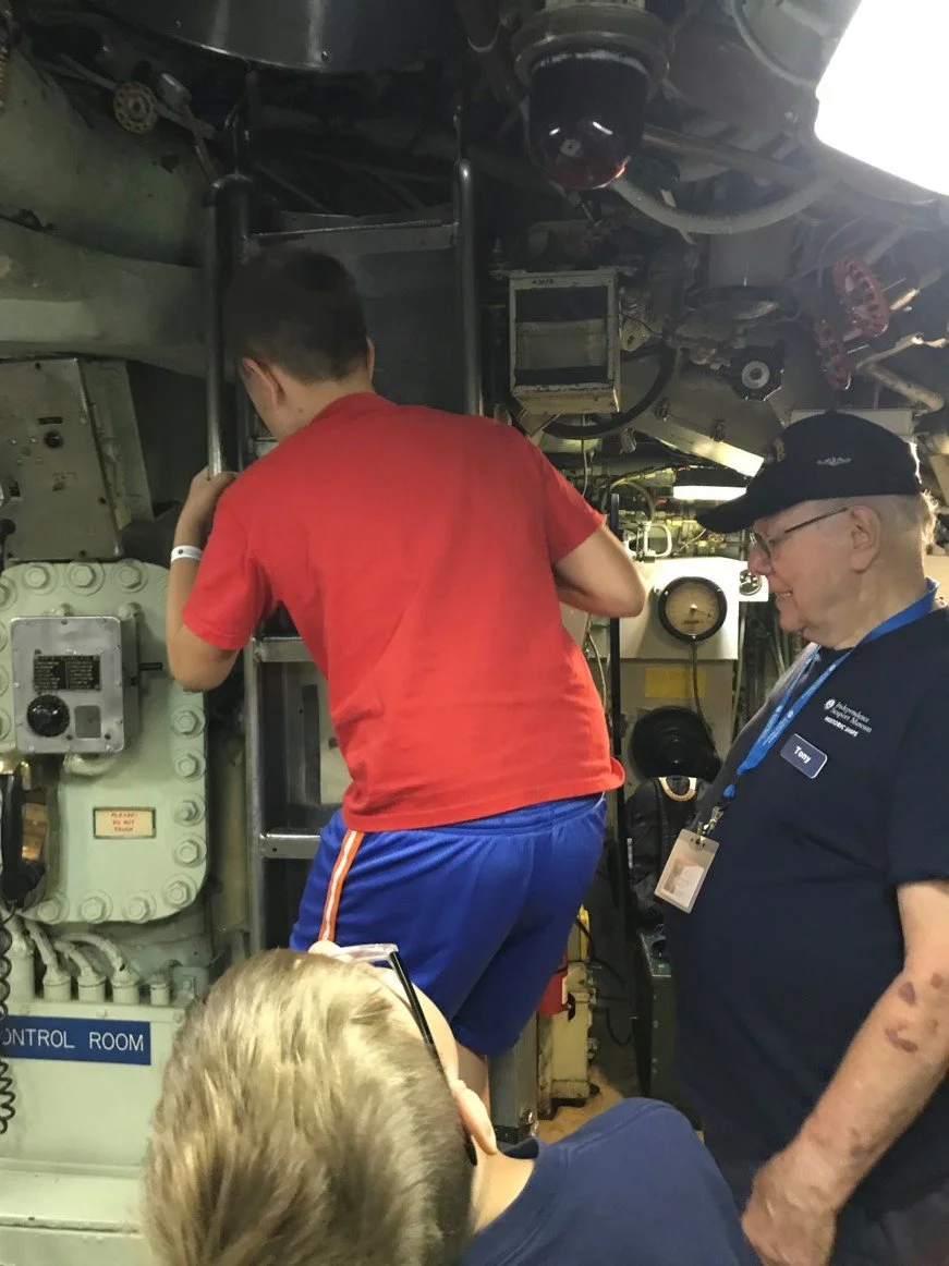 Three people working inside a submarine control room. One person is climbing a ladder, another is standing and watching, and a third is kneeling with glasses, possibly taking notes.