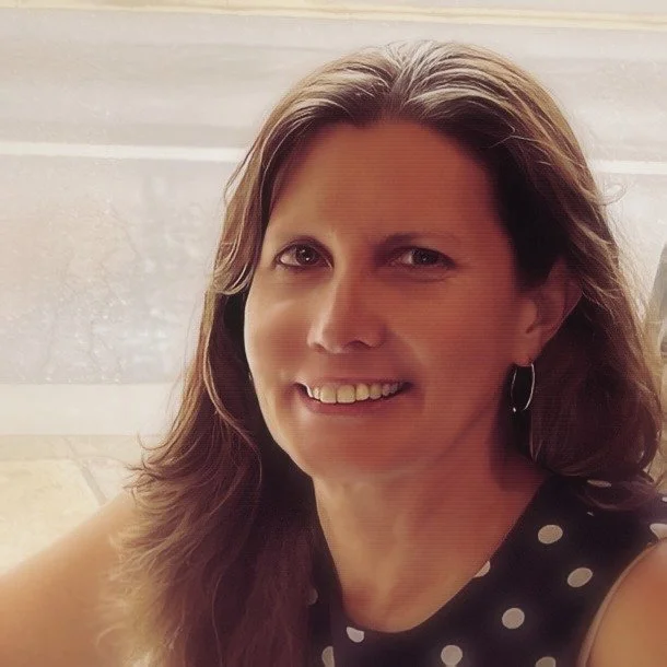 Woman with long brown hair smiling and winking, wearing a black dress with white polka dots, sitting near a window.