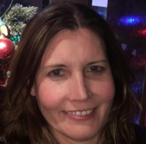 Woman with long brown hair smiling and winking, wearing a black dress with white polka dots, sitting near a window.