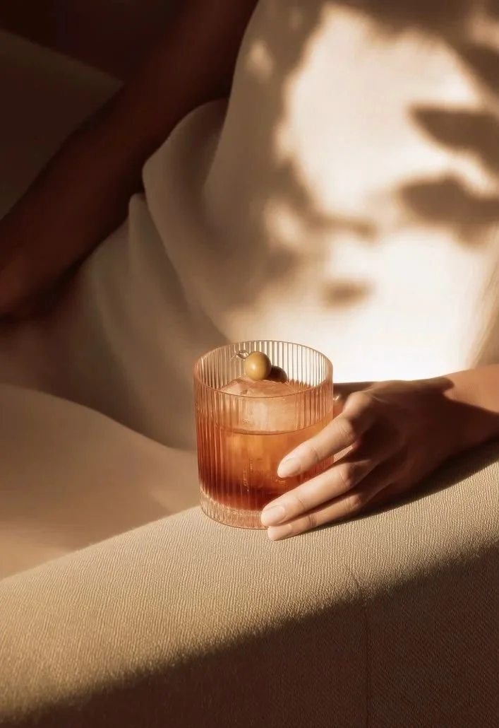 Close-up of a person's hand resting on a beige surface, holding a pink glass with a drink topped with a gold ball garnish, with soft lighting and shadow play.