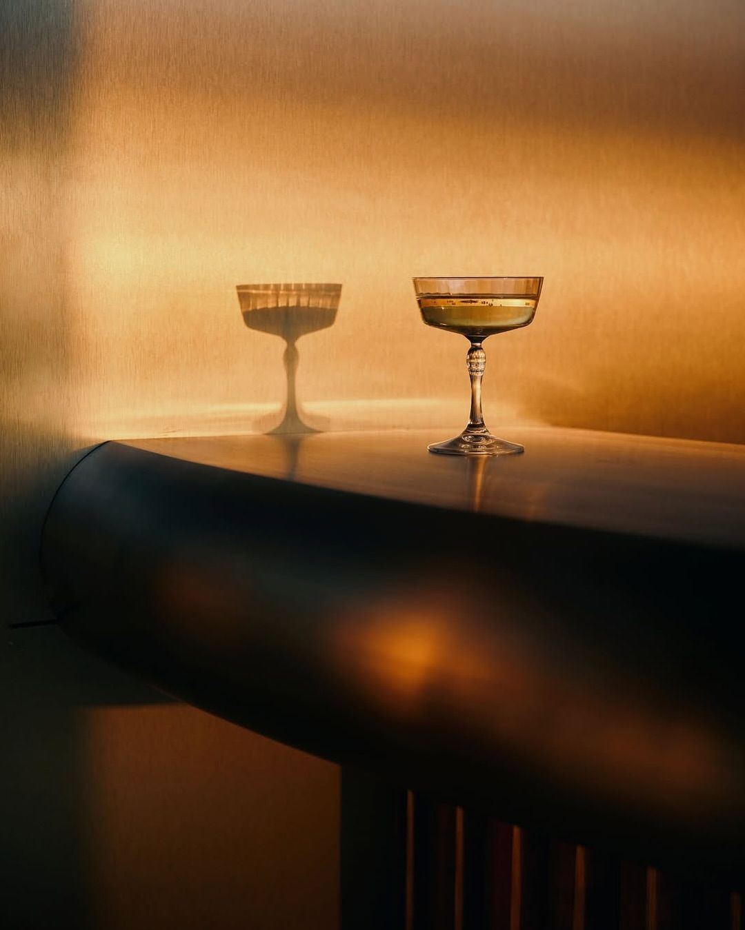 A cocktail glass with a golden drink sitting on a dark surface, casting a shadow with a cup-shaped outline on a beige wall.