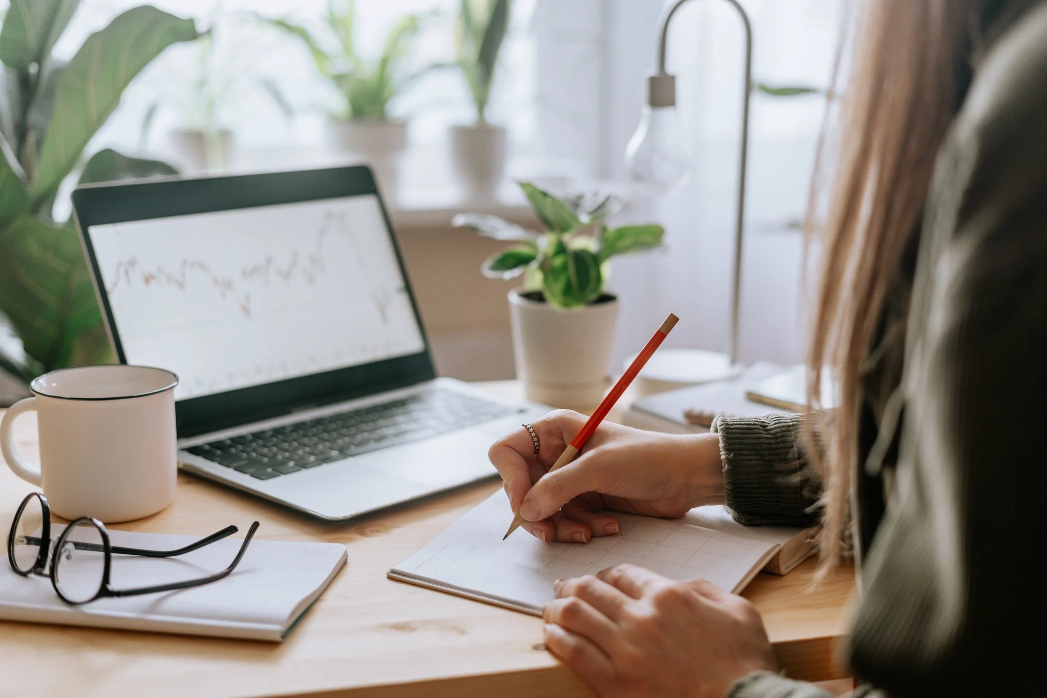 Person taking notes at a desk with a laptop, a coffee mug, and plants in the background.