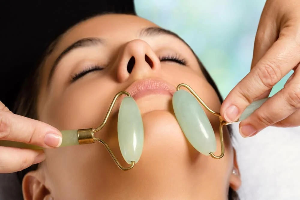 A woman receiving a facial massage with jade rollers on her face at a spa.