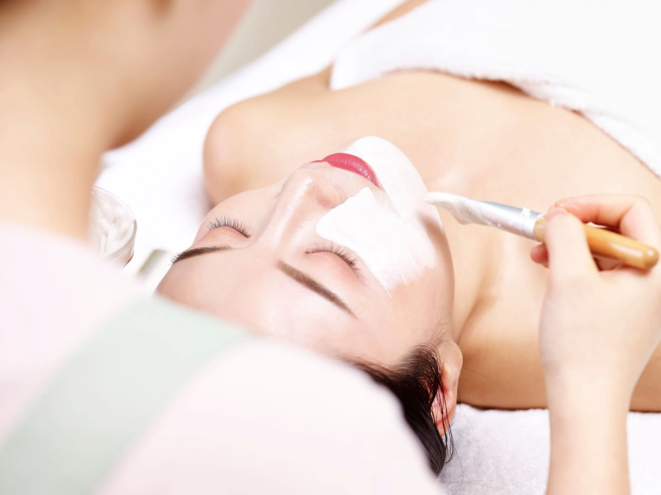A woman lying down receiving a facial treatment. A skincare specialist applies a white facial mask using a brush.