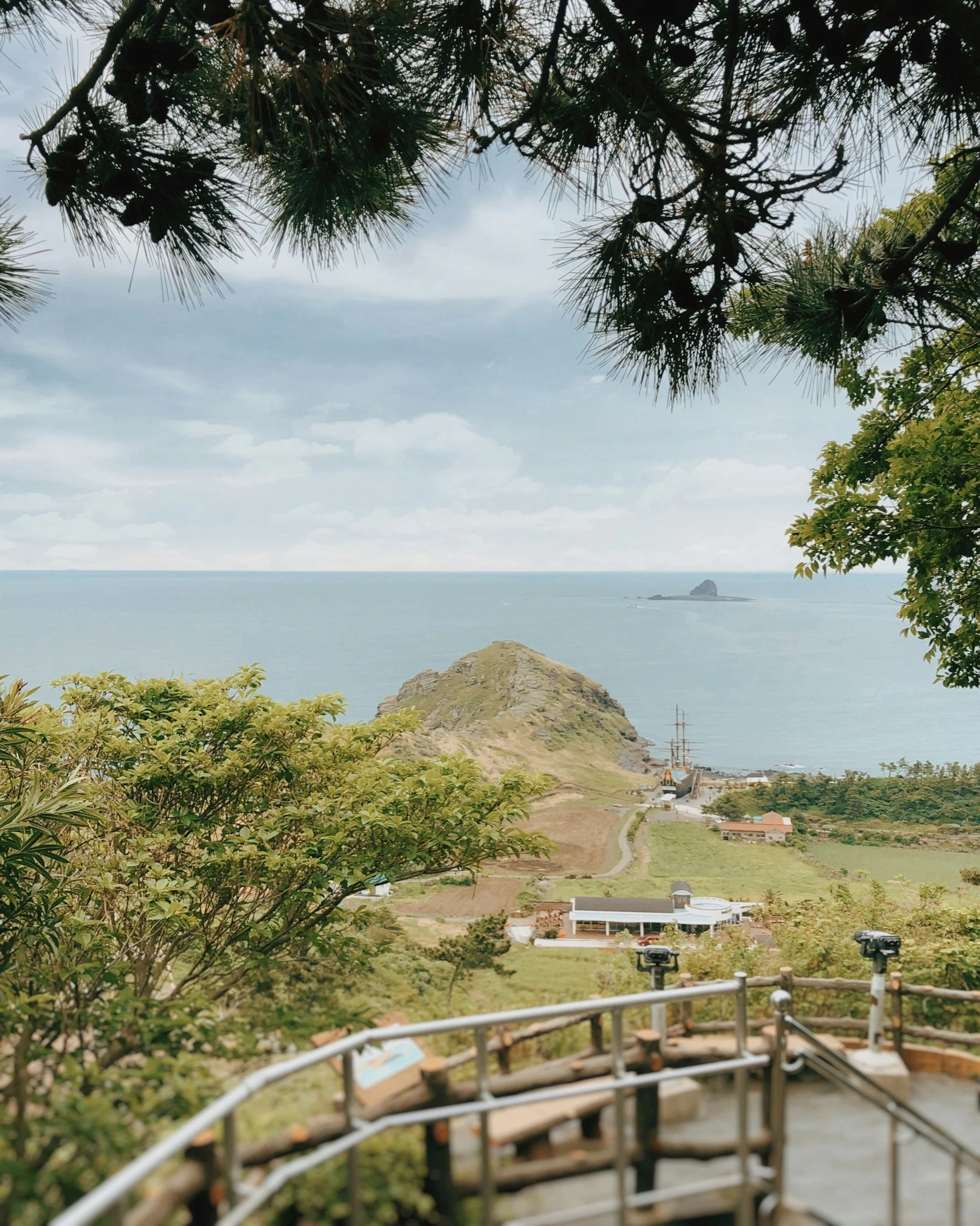 Scenic view of a coastal landscape with a small mountain or hill near the ocean, viewed from a lookout area with trees and a railing in the foreground.