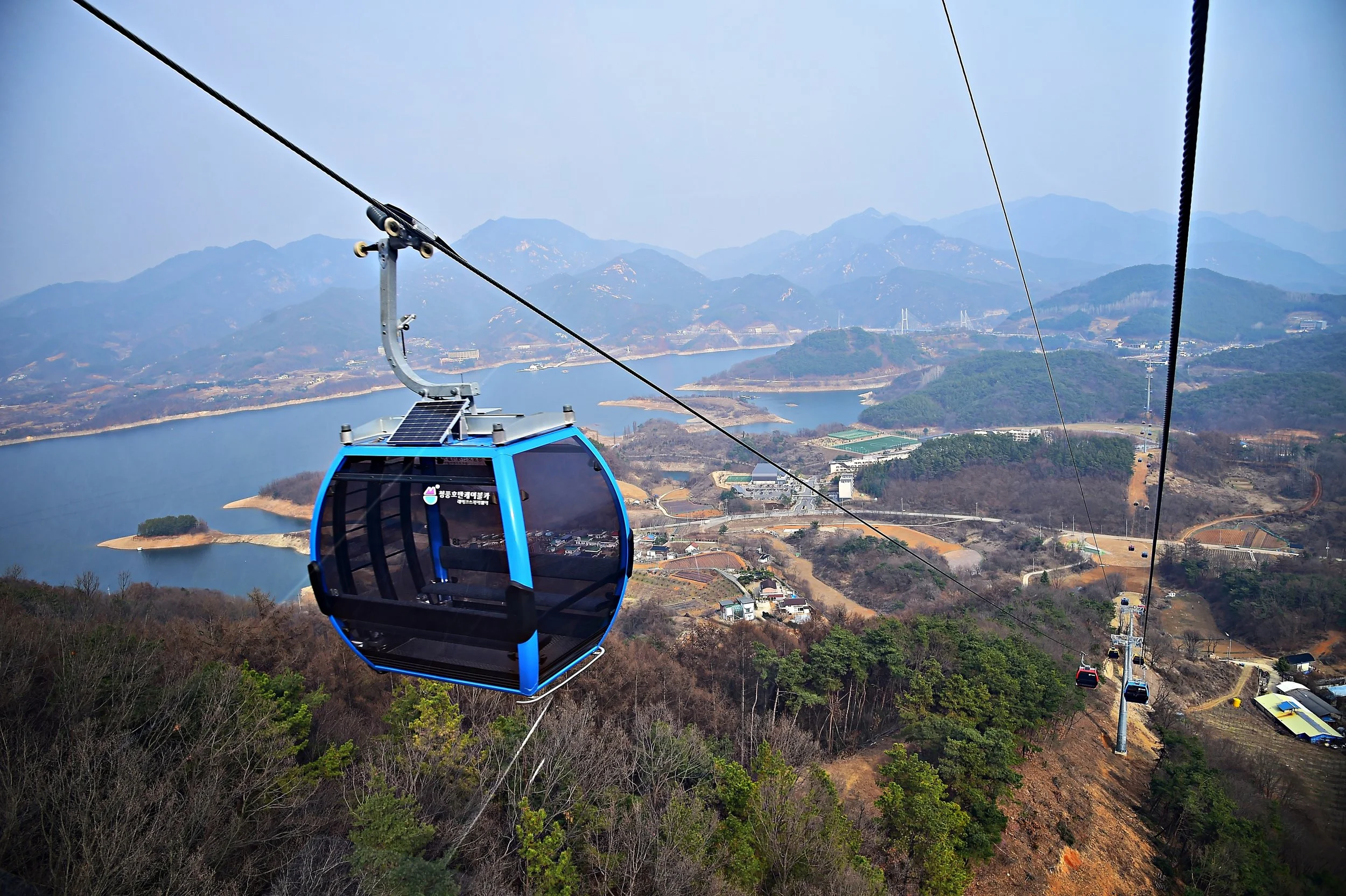 A blue gondola cabin on a cable car system flying over a landscape with a lake, trees, and distant mountains.