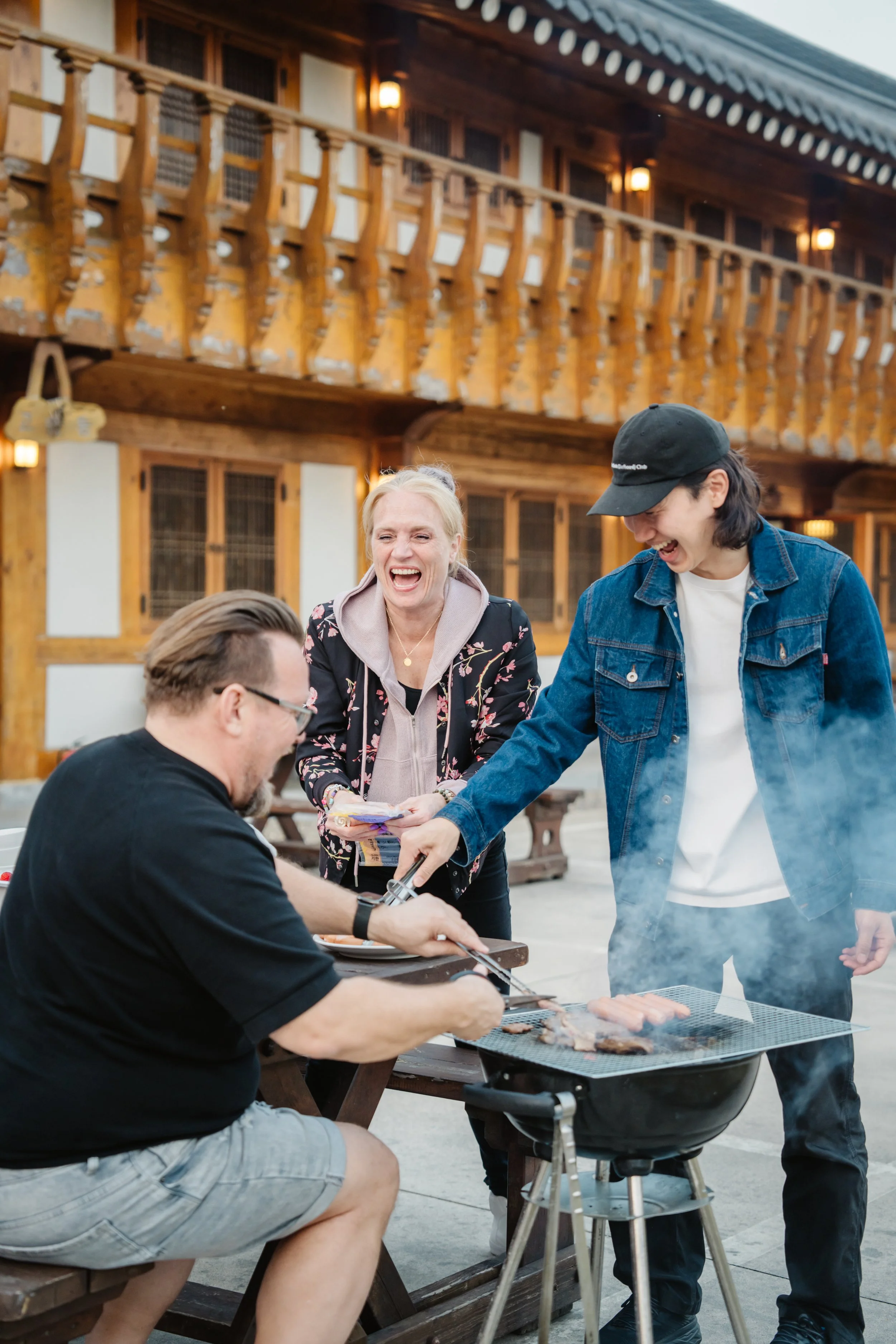 Three people laughing and enjoying a barbecue outside a wooden lodge-style building, with smoke rising from the grill.