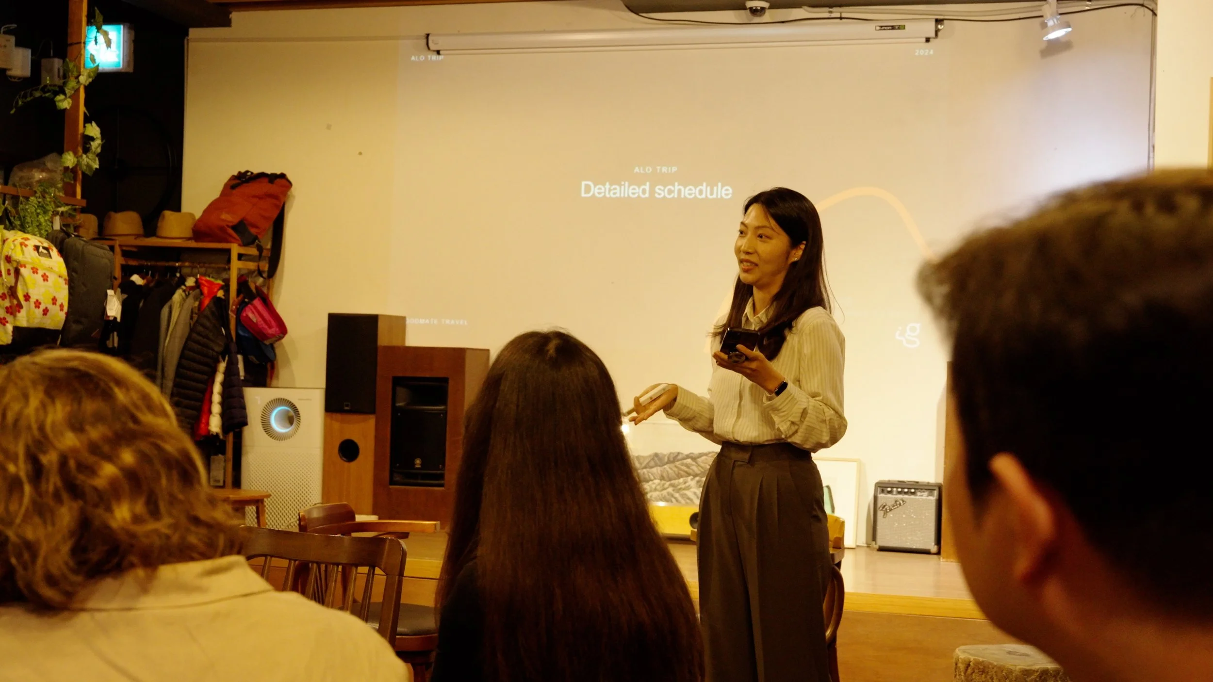 A woman giving a presentation in a room with audience members seated and a projector screen displaying text. The room has shelves with bags and hats on the left side.