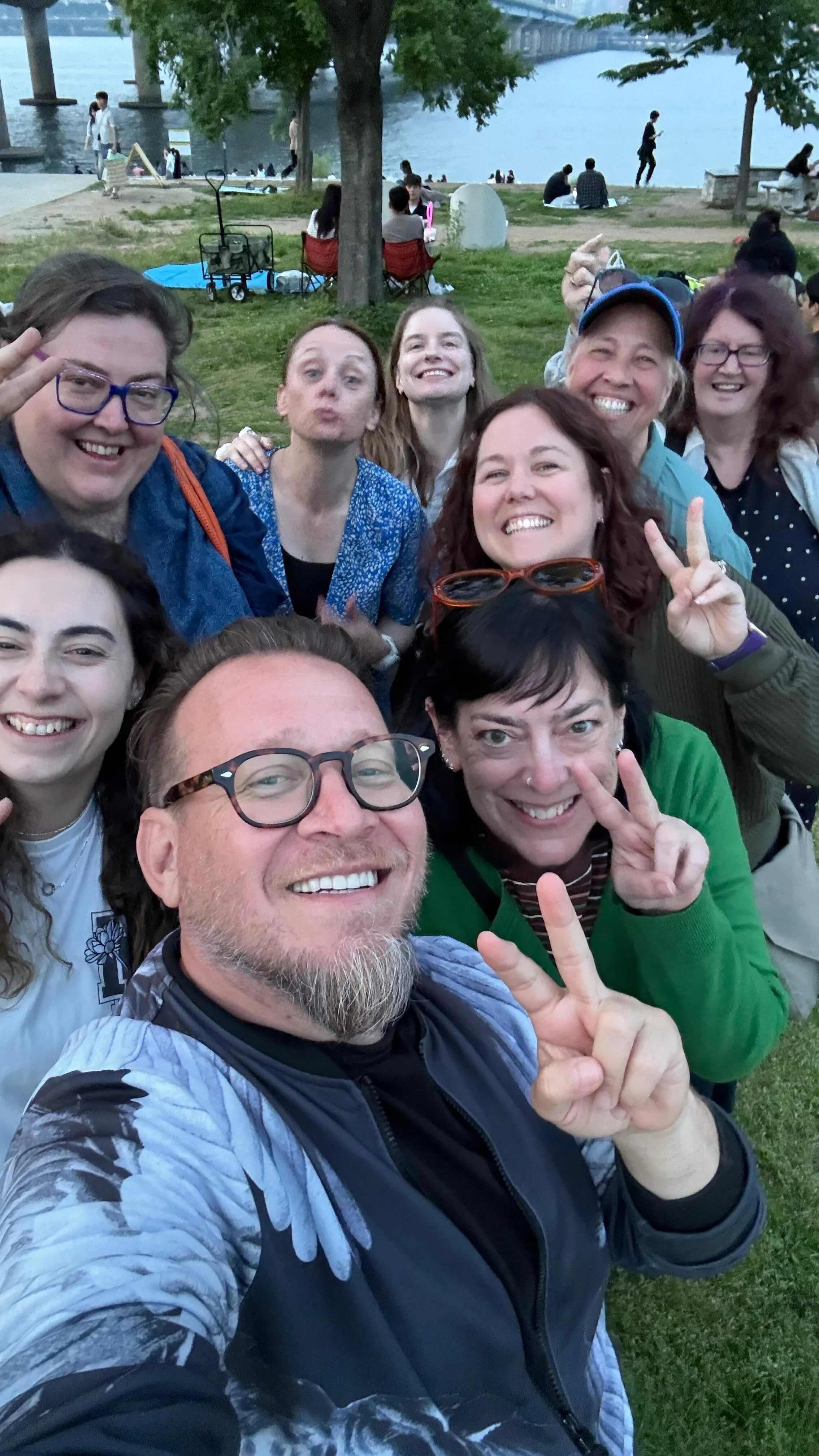 Group of people taking a selfie by a river in a park, with trees and a bridge in the background, some making peace signs and smiling.