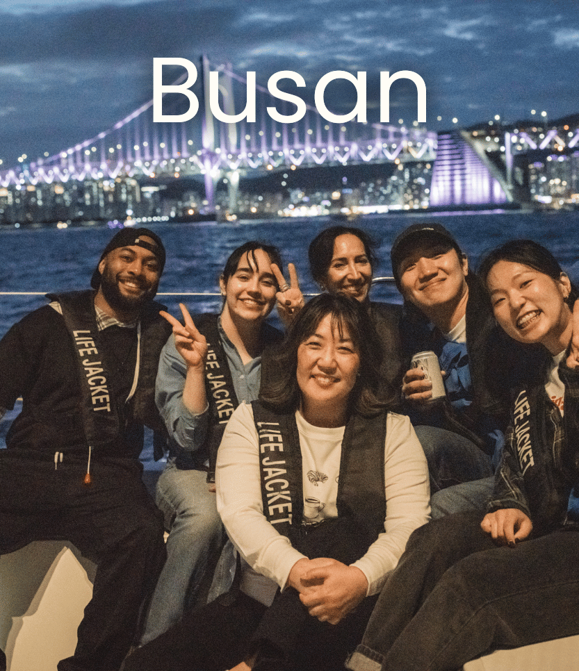 Group of six friends smiling and making peace signs on a boat at night in front of a lit-up bridge in Busan, South Korea.