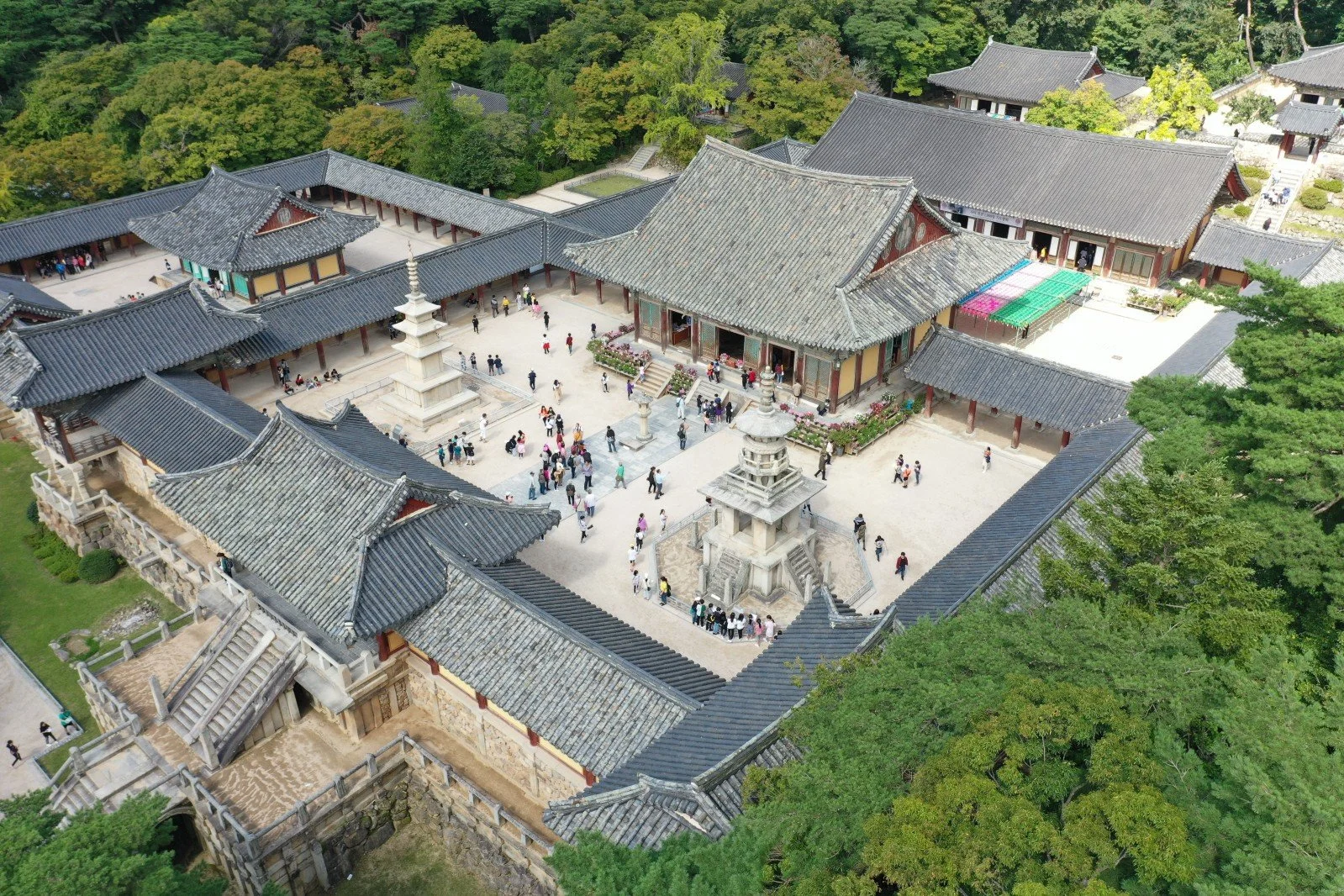 An aerial view of a traditional Asian temple complex with gray tiled roofs, stone pagodas, and visitors walking in the courtyard surrounded by lush green trees.