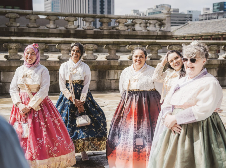 Five women wearing traditional Korean hanbok dresses standing outdoors on a stone terrace with a cityscape in the background, smiling and posing for the photo.