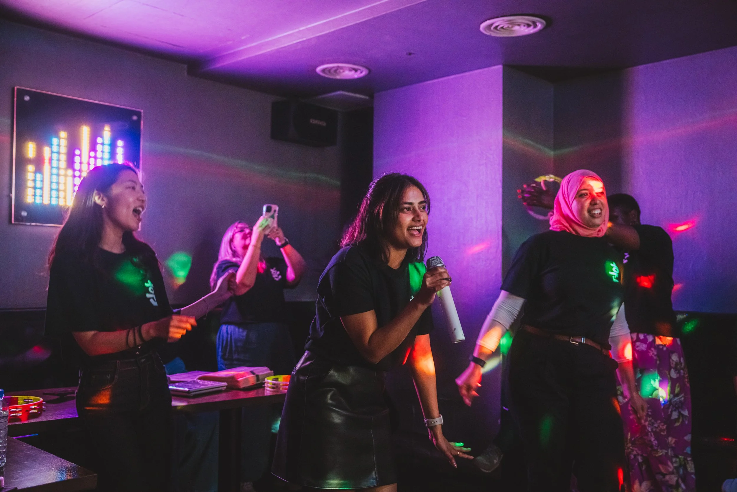 Group of women singing and dancing at a karaoke party with colorful lights and a DJ setup.
