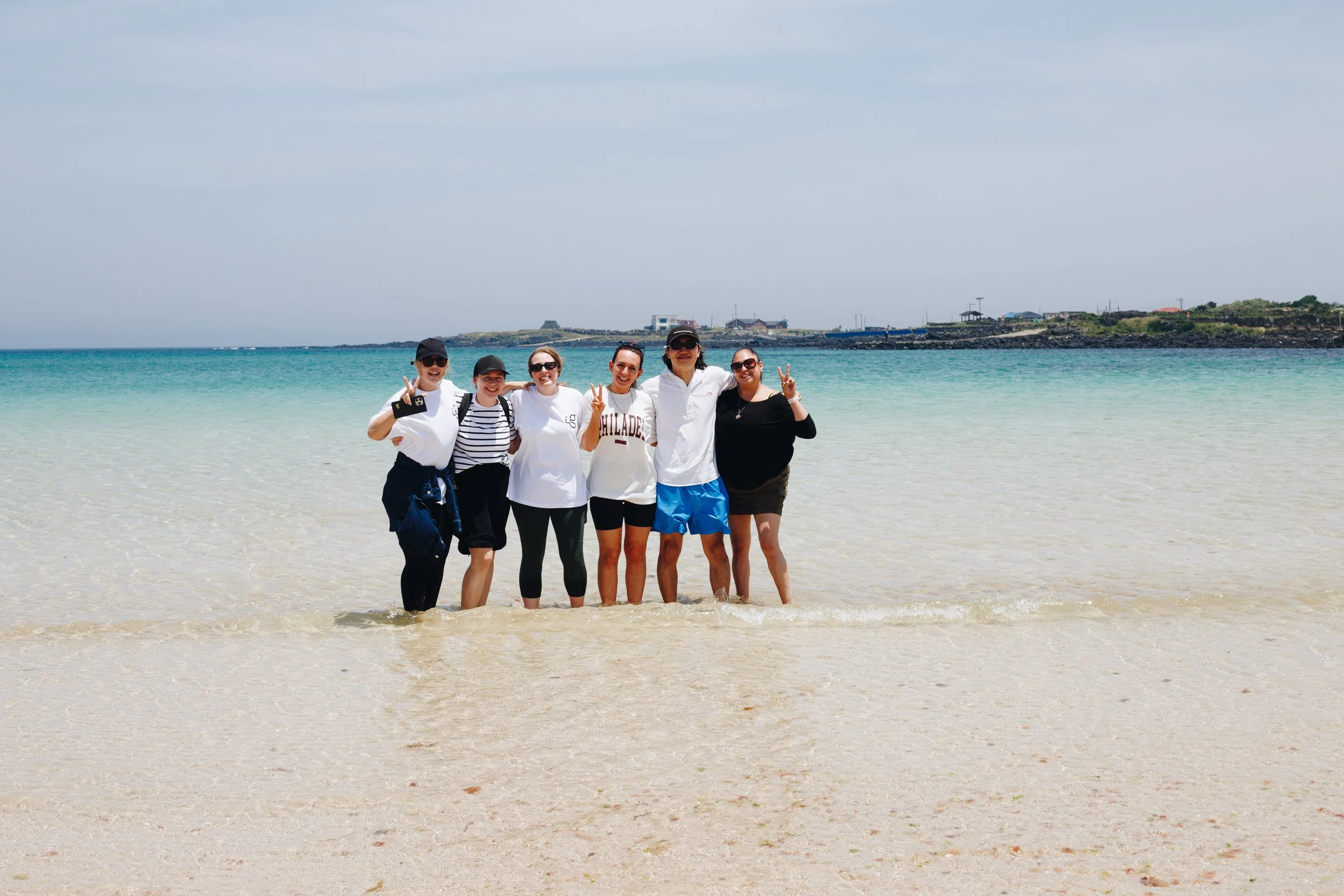 Six friends standing in shallow ocean water at a beach, smiling and making peace signs.