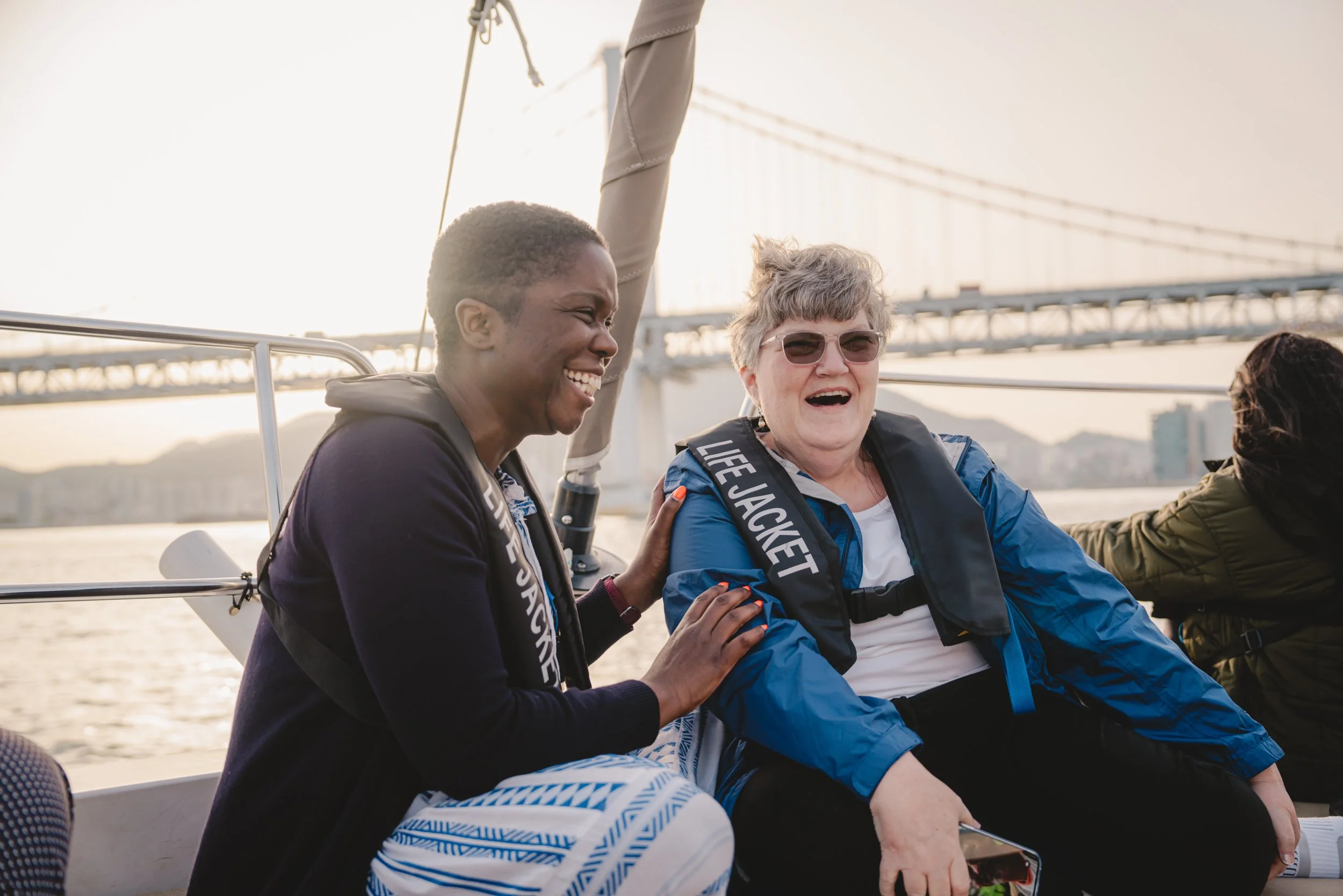 Two women sitting on a boat, laughing and enjoying each other's company, with a bridge in the background.