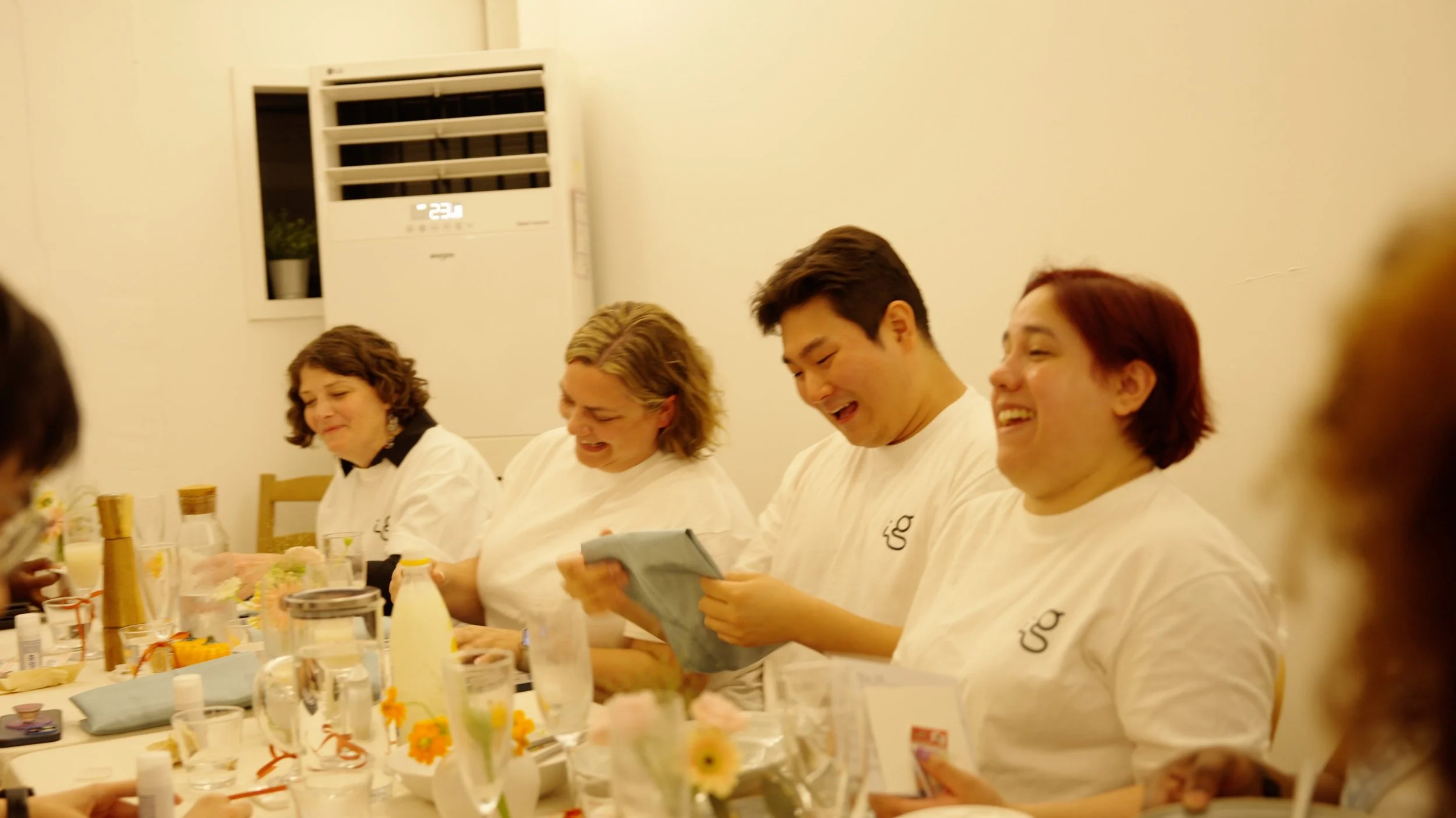 Group of people sitting at a long table, smiling and laughing during a gathering or event. The table is decorated with flowers, glasses, and bottles, and everyone appears to be enjoying themselves.
