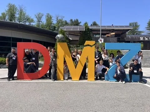 Group of people posing behind large, colorful letters spelling DMI, outdoors with trees and a modern building in the background.