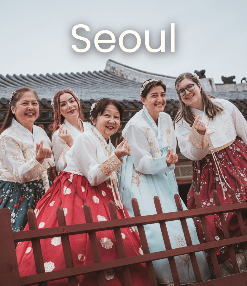 Five women wearing traditional Korean hanbok clothing, standing in front of a traditional Korean building with a tiled roof, smiling and making hand gestures, with the word 'Seoul' displayed above.