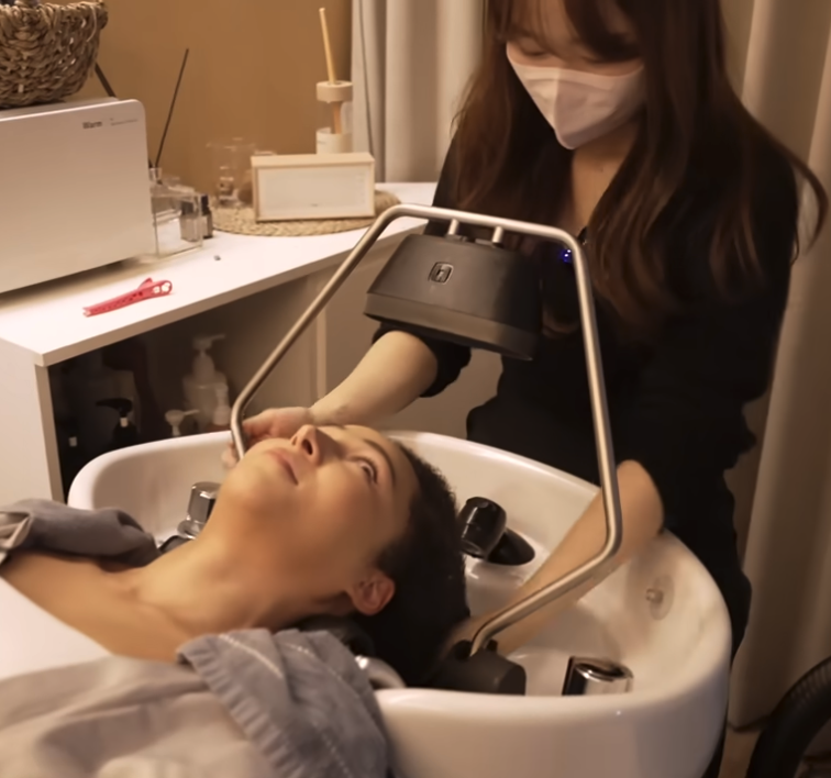 Woman in a white mask washing a woman's hair at a salon sink.
