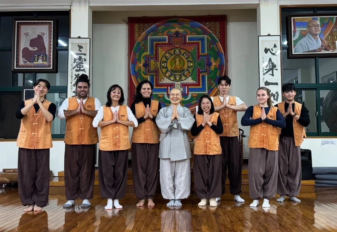 Group of nine people standing in a religious or cultural setting, wearing traditional clothing with their hands pressed together in a prayer gesture, in front of colorful wall art and framed portraits.