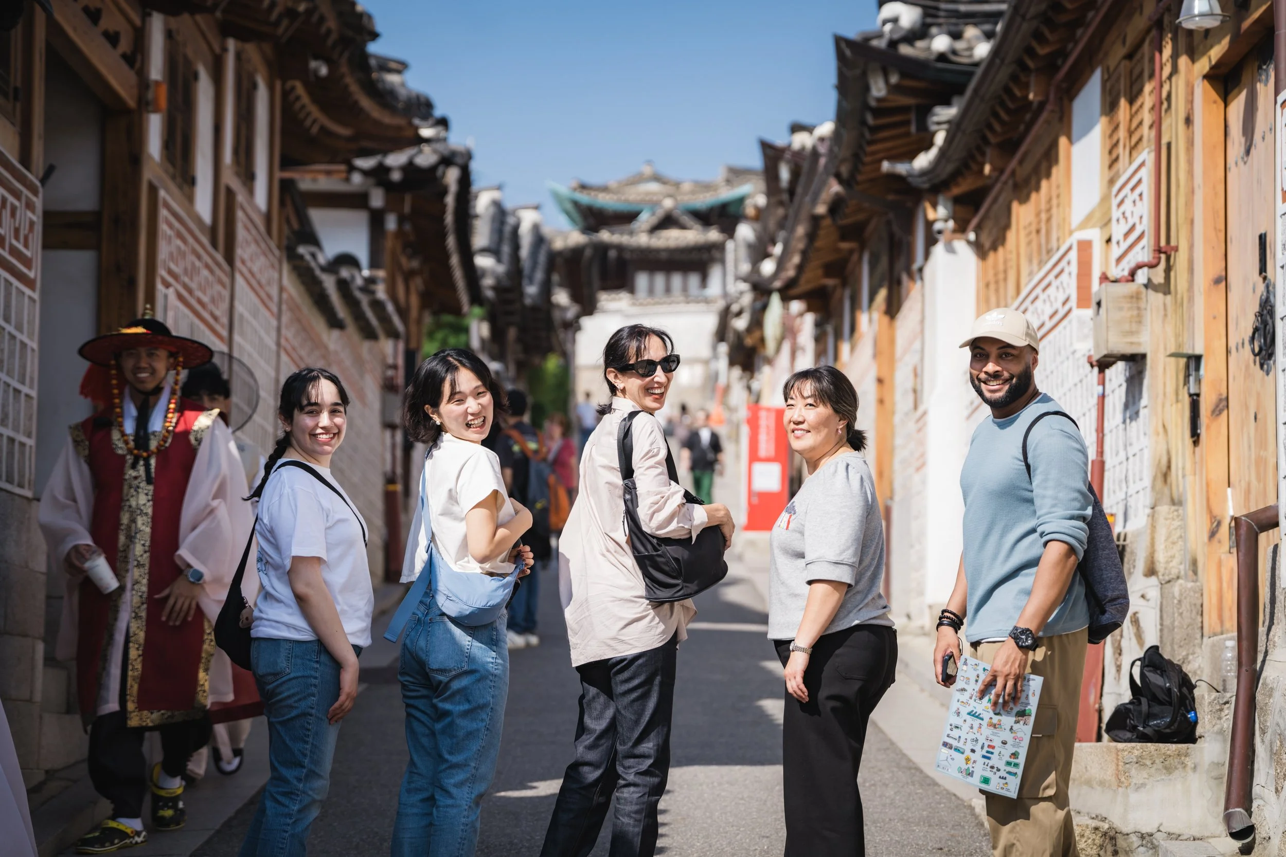 Group of diverse tourists smiling and exploring a traditional Korean village street with ornate wooden buildings under a clear blue sky.