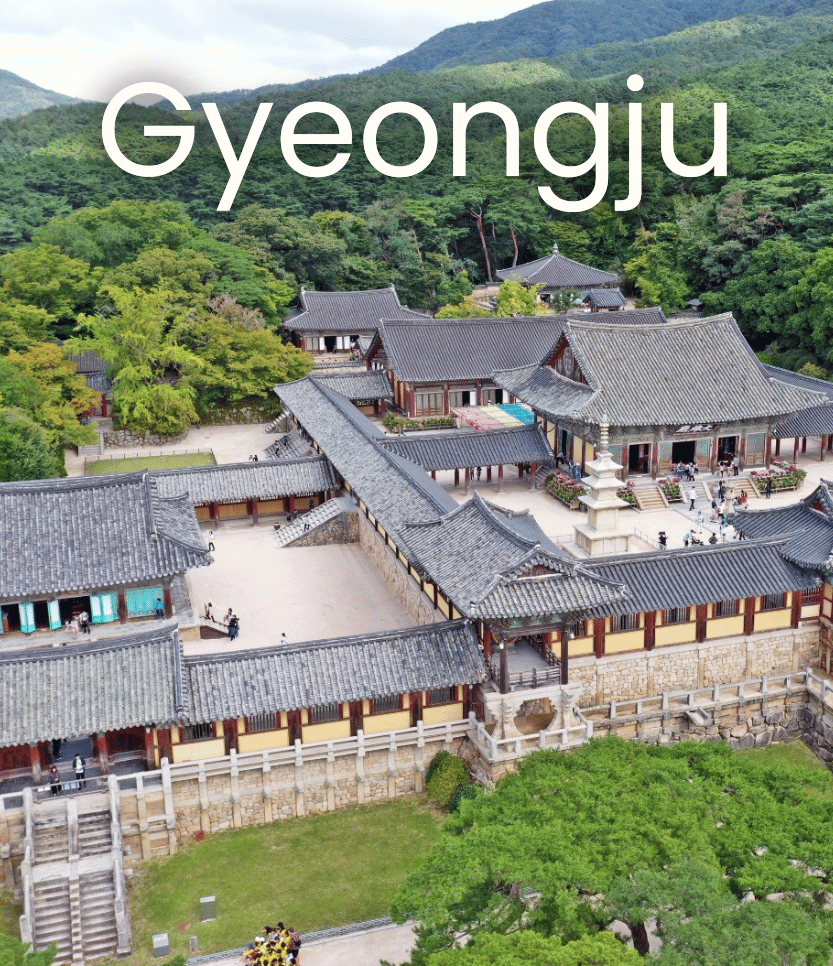 A traditional Korean temple complex surrounded by green trees and mountains, with tiled roofs and stone walls.