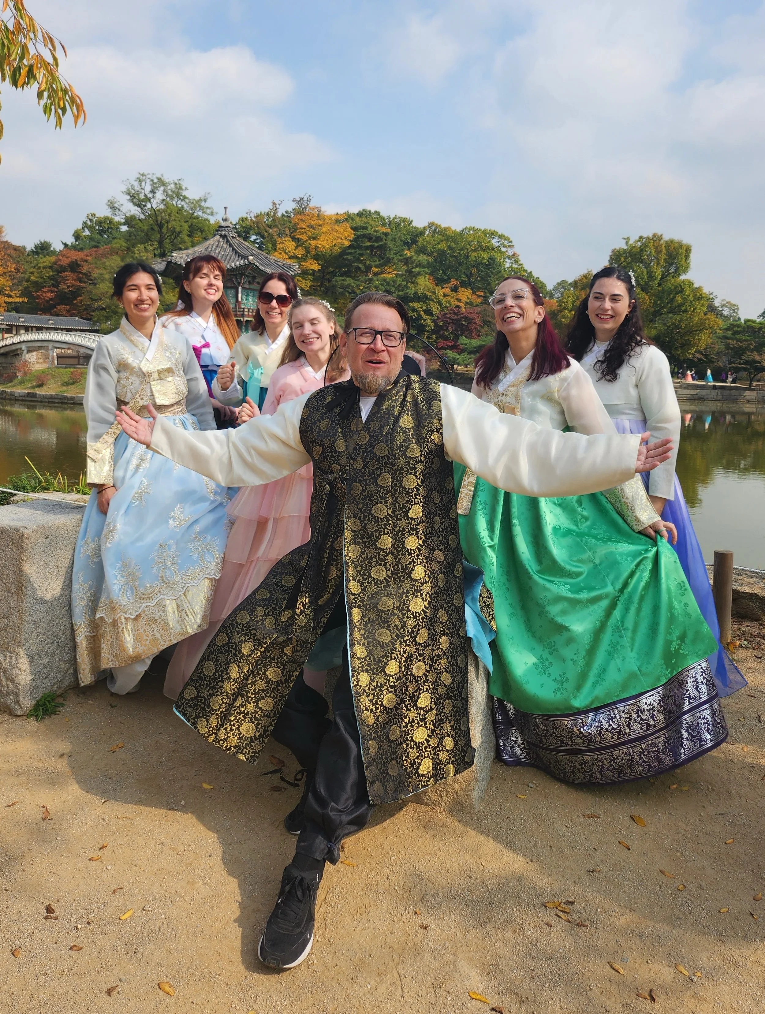 A man with glasses and a beard, wearing a traditional Korean hanbok, stands with arms outstretched in front of six women dressed in colorful hanboks near a lake, with trees and traditional Korean architecture in the background.