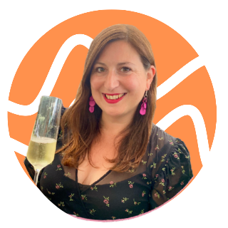 A woman with shoulder-length brown hair, red lipstick, wearing purple earrings and a black floral dress, smiling and holding a glass of champagne.