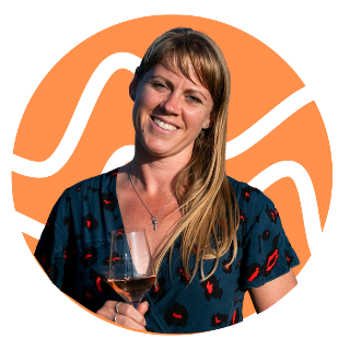 A woman with light brown hair, wearing a blue dress with red and black patterns, holding a glass of rosé wine, smiling, with a pink circle background.