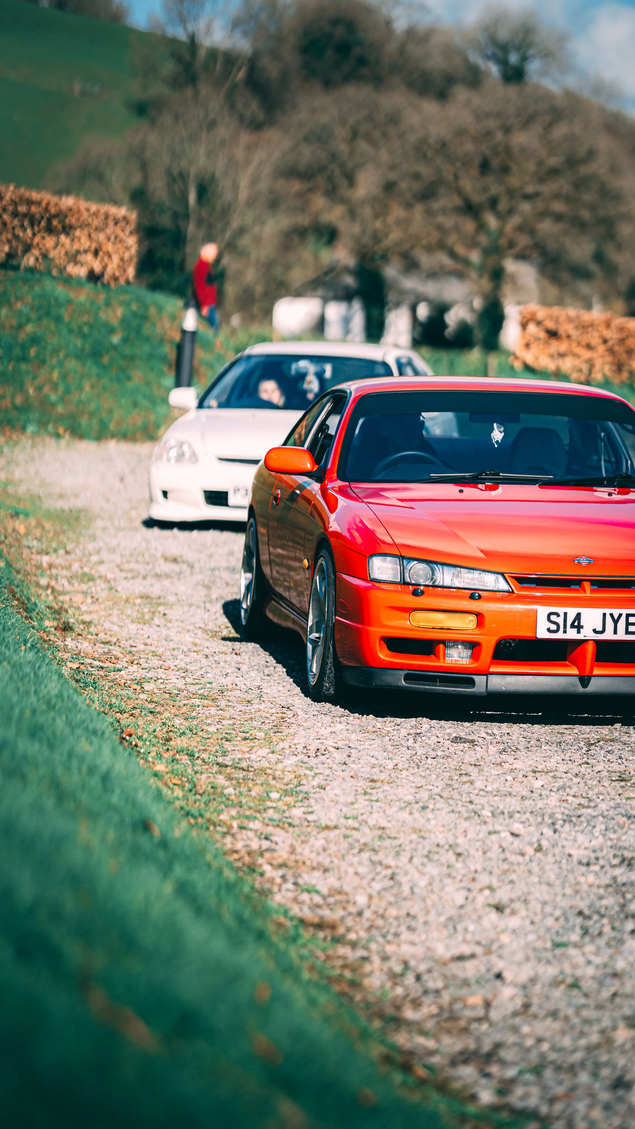 A red sports car parked on a gravel pathway with a white car behind it, and a person standing near a bush in a rural area with trees and hills in the background.