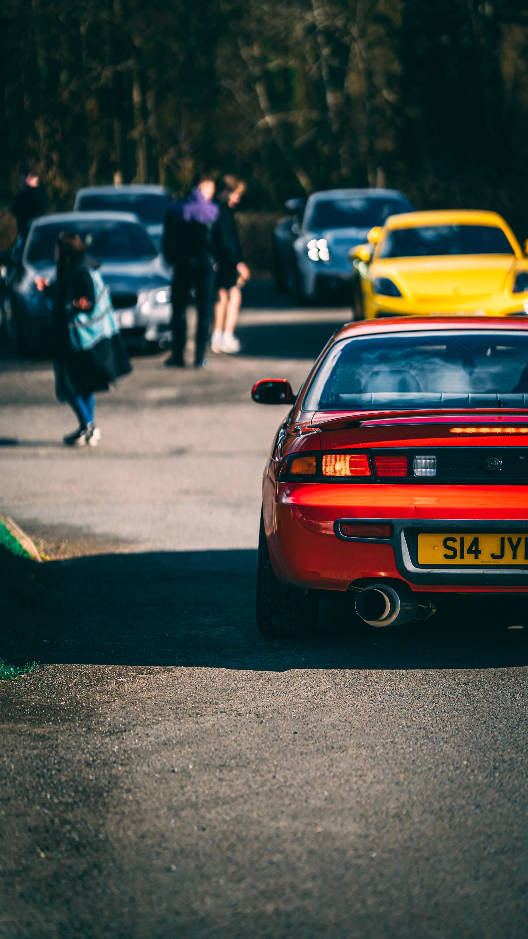 Red sports car parked on the side of a road with a group of people and several parked cars in the background, with trees in the distance.
