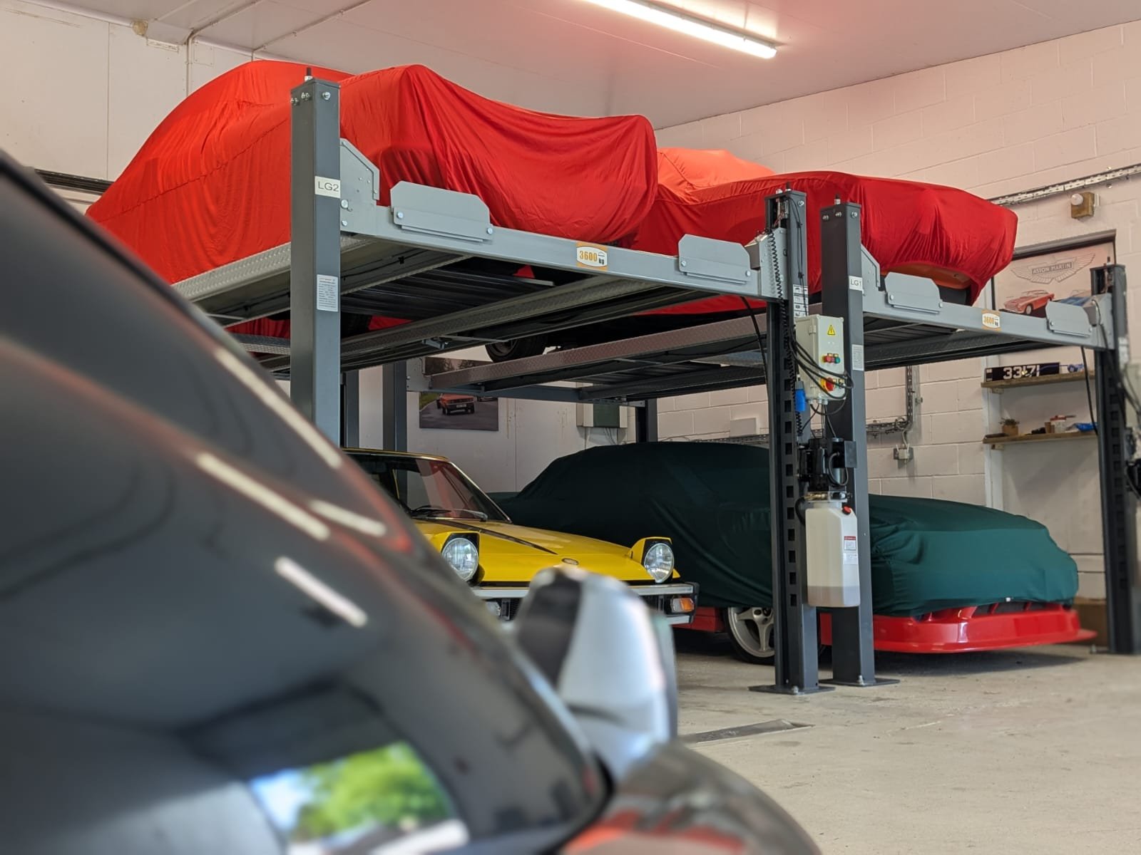 Three cars stored on a hydraulic lift in a garage, two with covers and one partially visible in front.