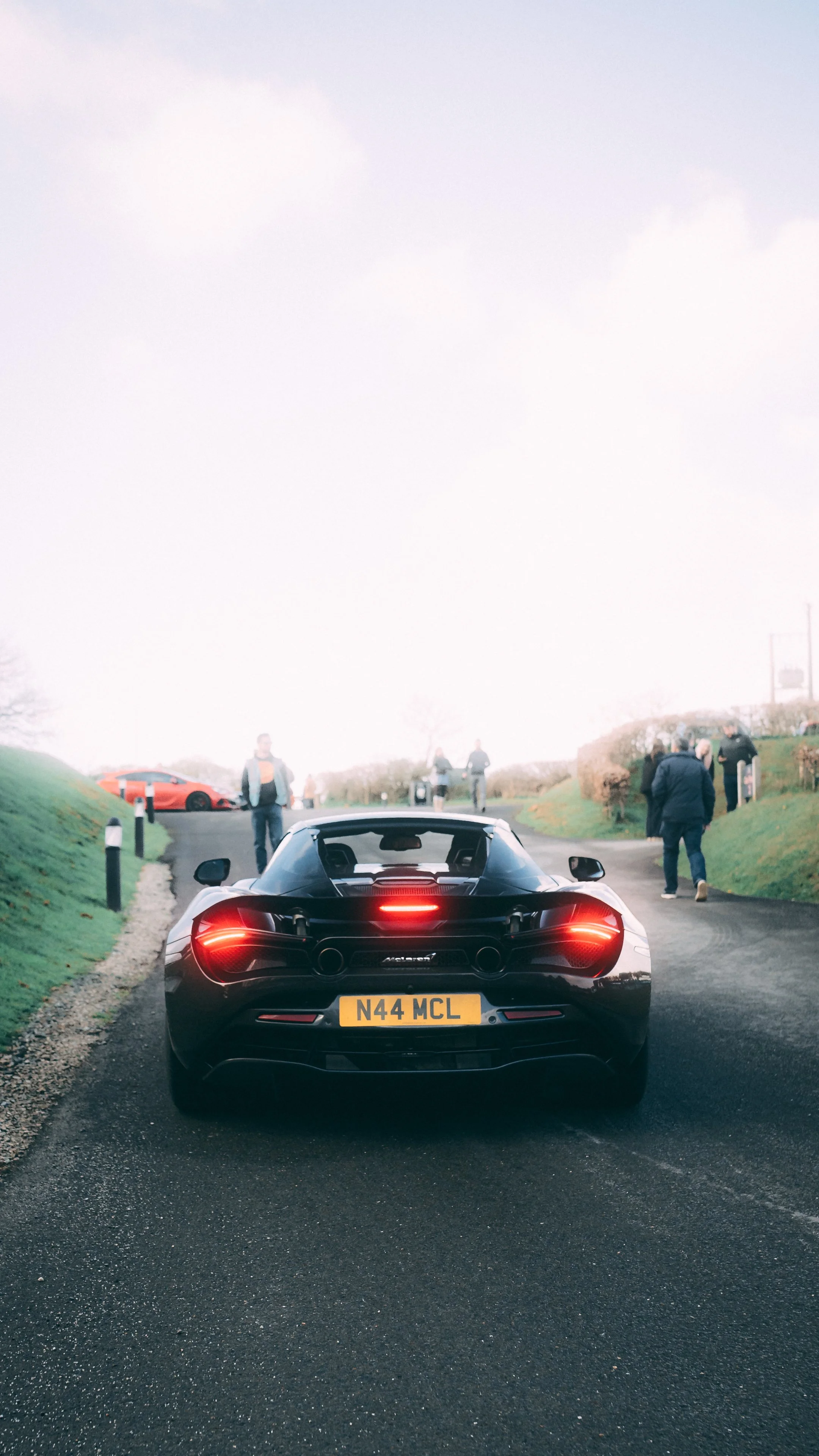 Black sports car with a rear spoiler and distinctive rear lights, driving on a wet road with a group of people standing and walking nearby on a cloudy day.