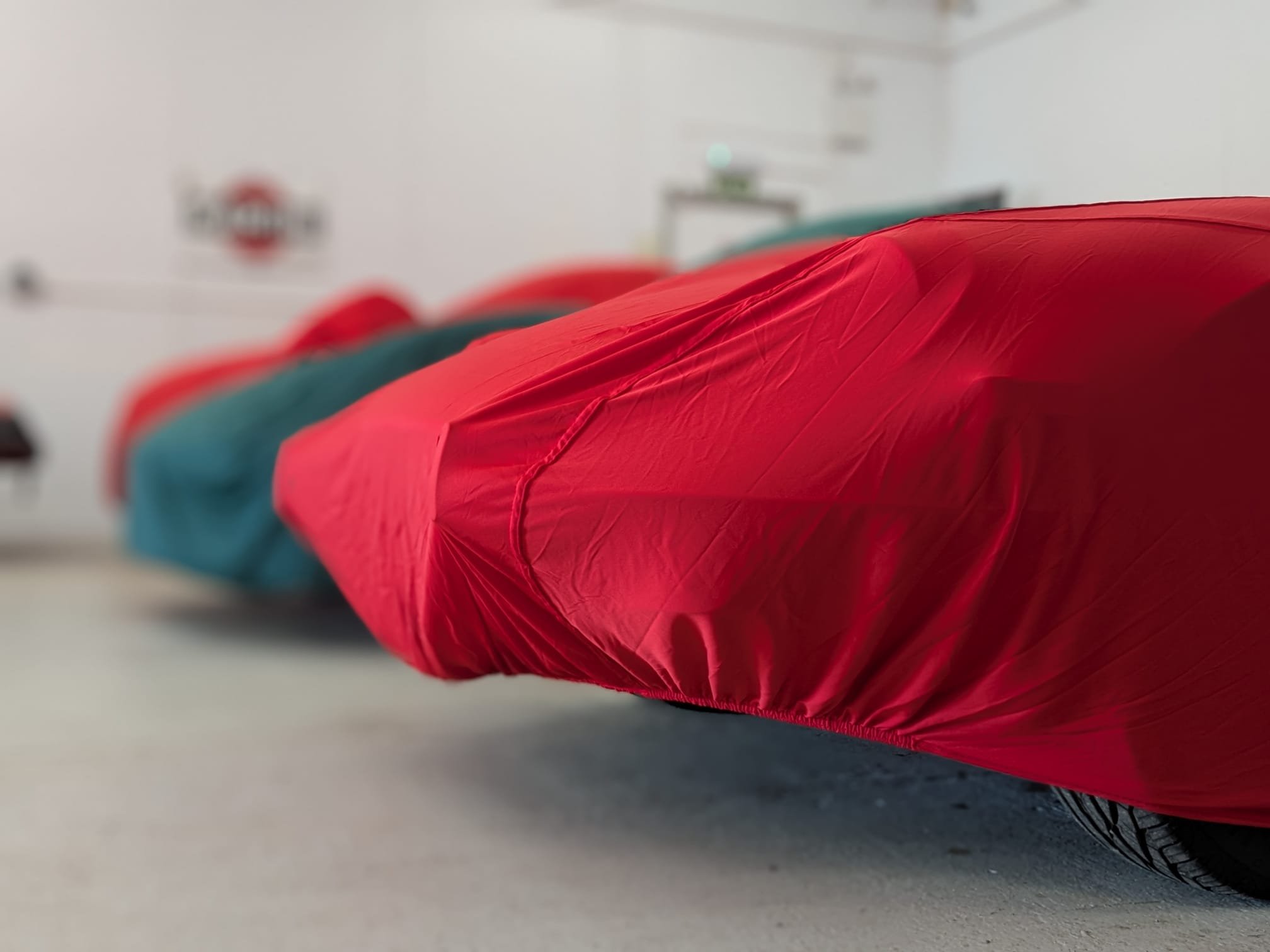 Close-up of a row of cars covered with colorful car covers, red and teal, inside a garage or showroom.