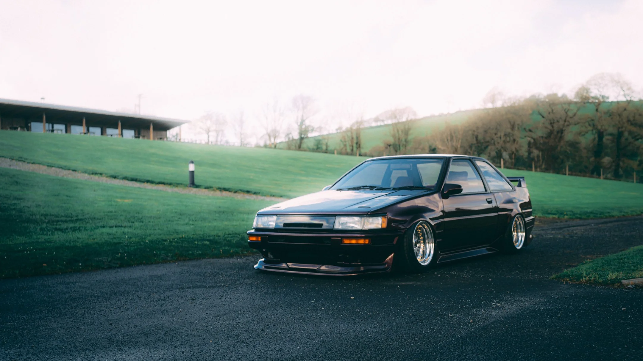 A black vintage sports car parked on a paved area near a grassy hill with trees and a building in the background.