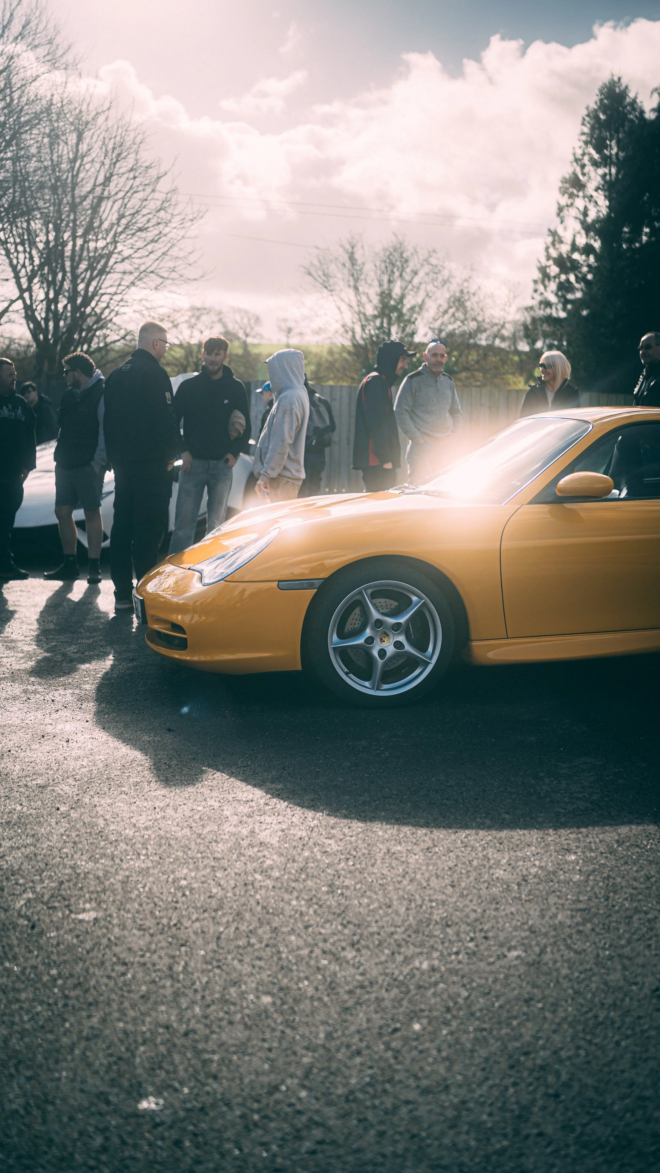 A group of people standing outdoors near a yellow sports car, with a blue sky and trees in the background.