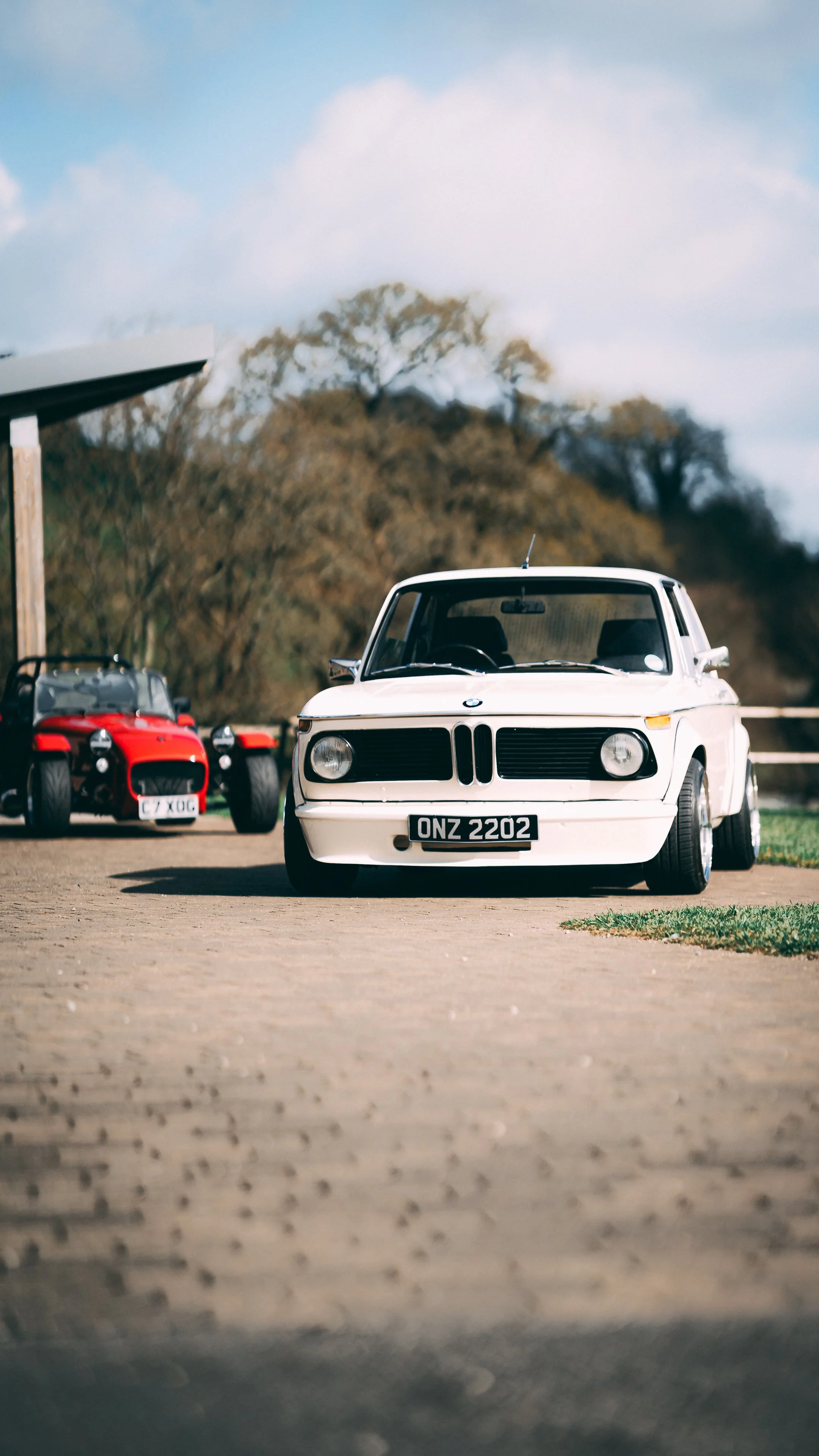 A white vintage car parked outdoors with a black front grille, round headlights, and a black license plate reading 'ONZ 2202', with a red sports car in the background and a scenic landscape of trees and a cloudy sky.