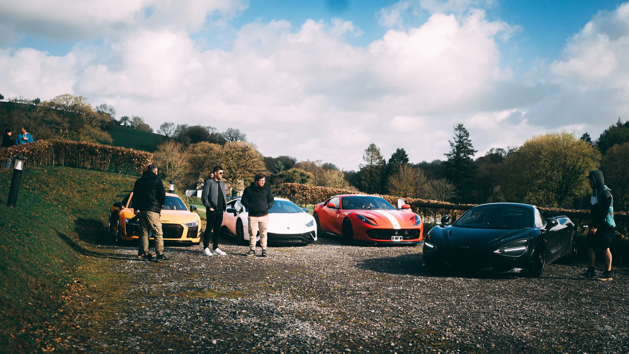 Group of people standing near luxury sports cars parked outdoors, with trees and a cloudy sky in the background.