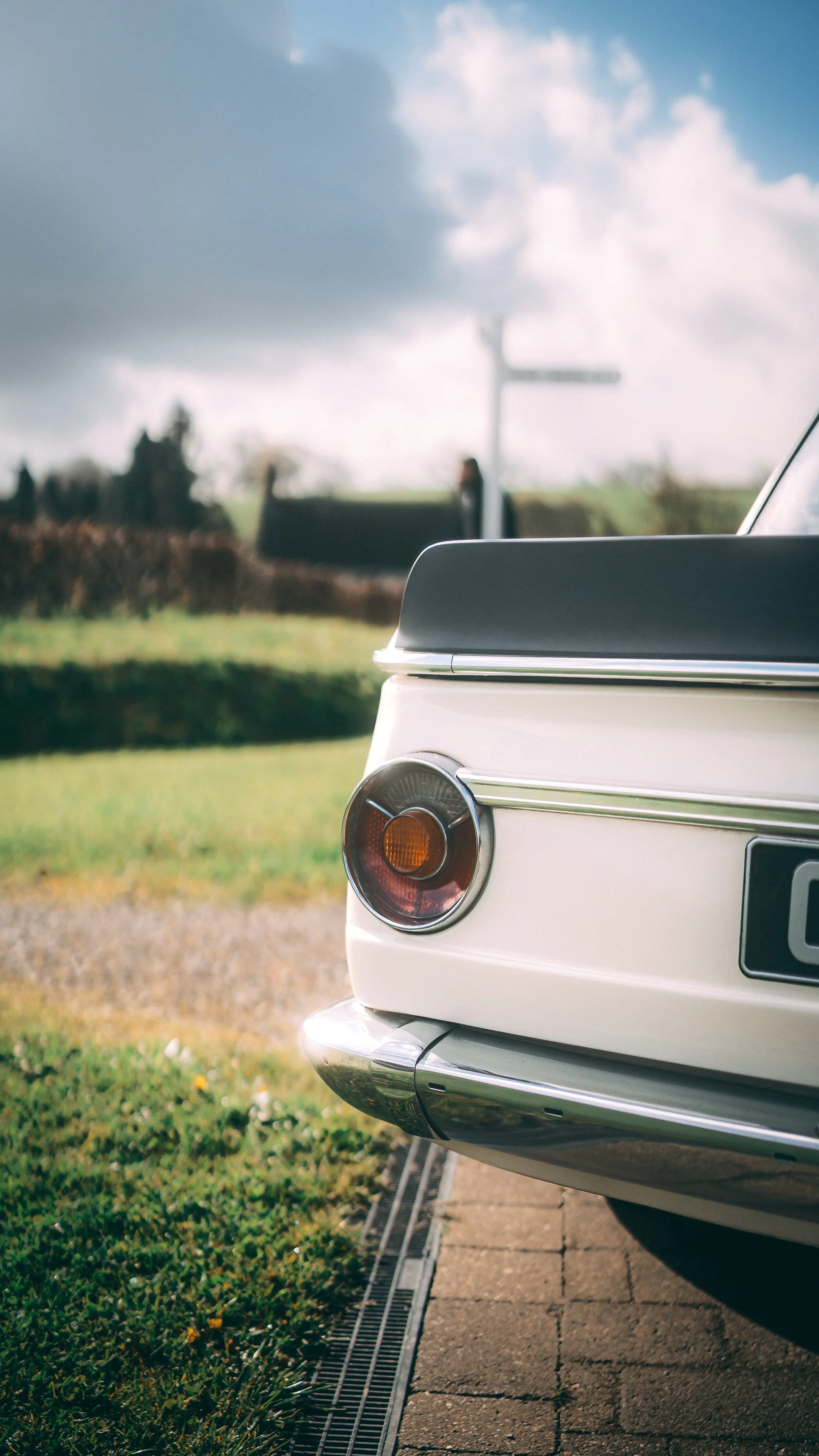 Close-up of the rear of a white vintage car with a round taillight and a black spoiler, parked on a brick driveway with a grassy area and a road in the background under a partly cloudy sky.