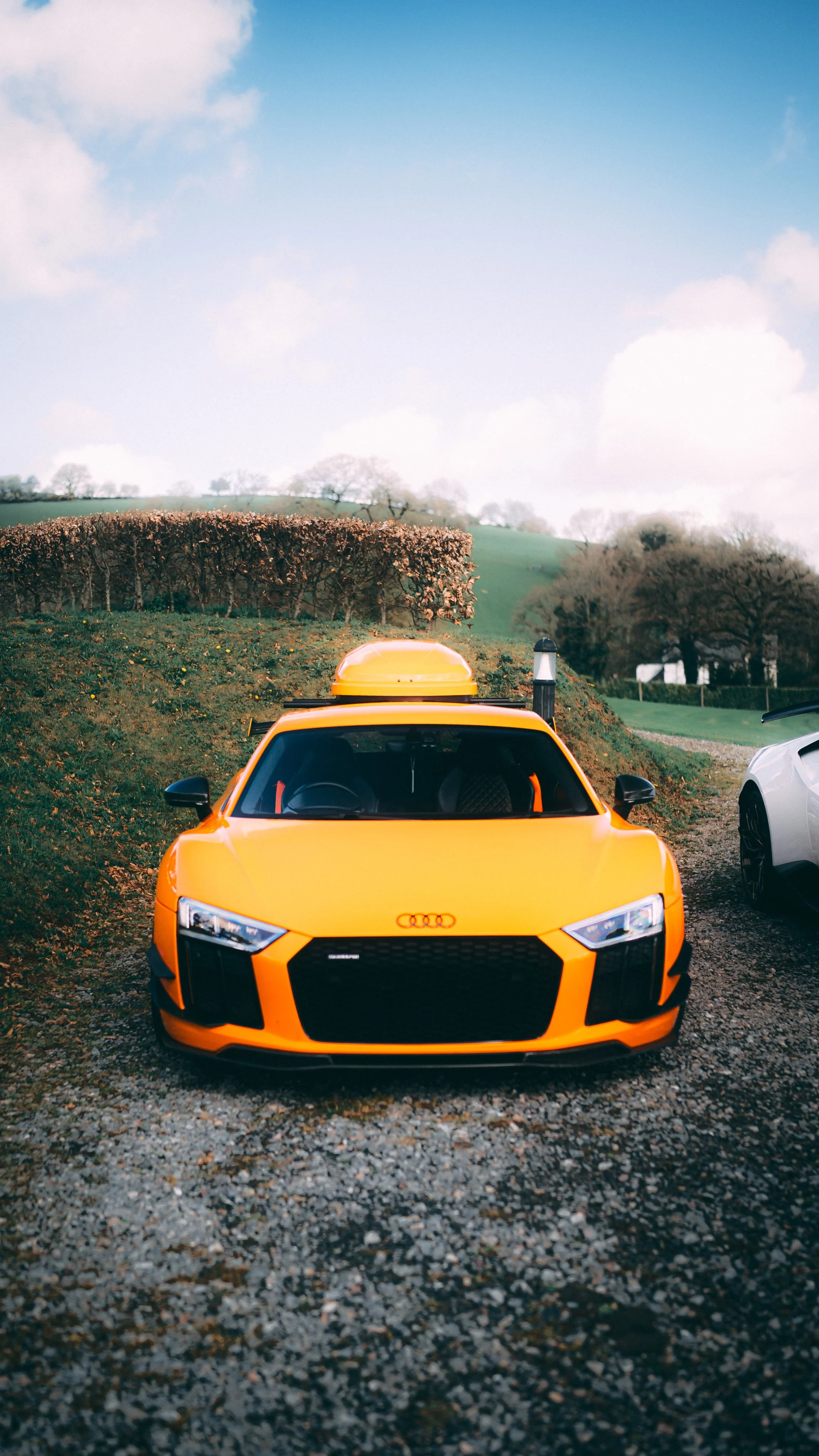 Front view of an orange Audi sports car parked on a gravel driveway with a landscaped yard and trees in the background, under a partly cloudy sky.