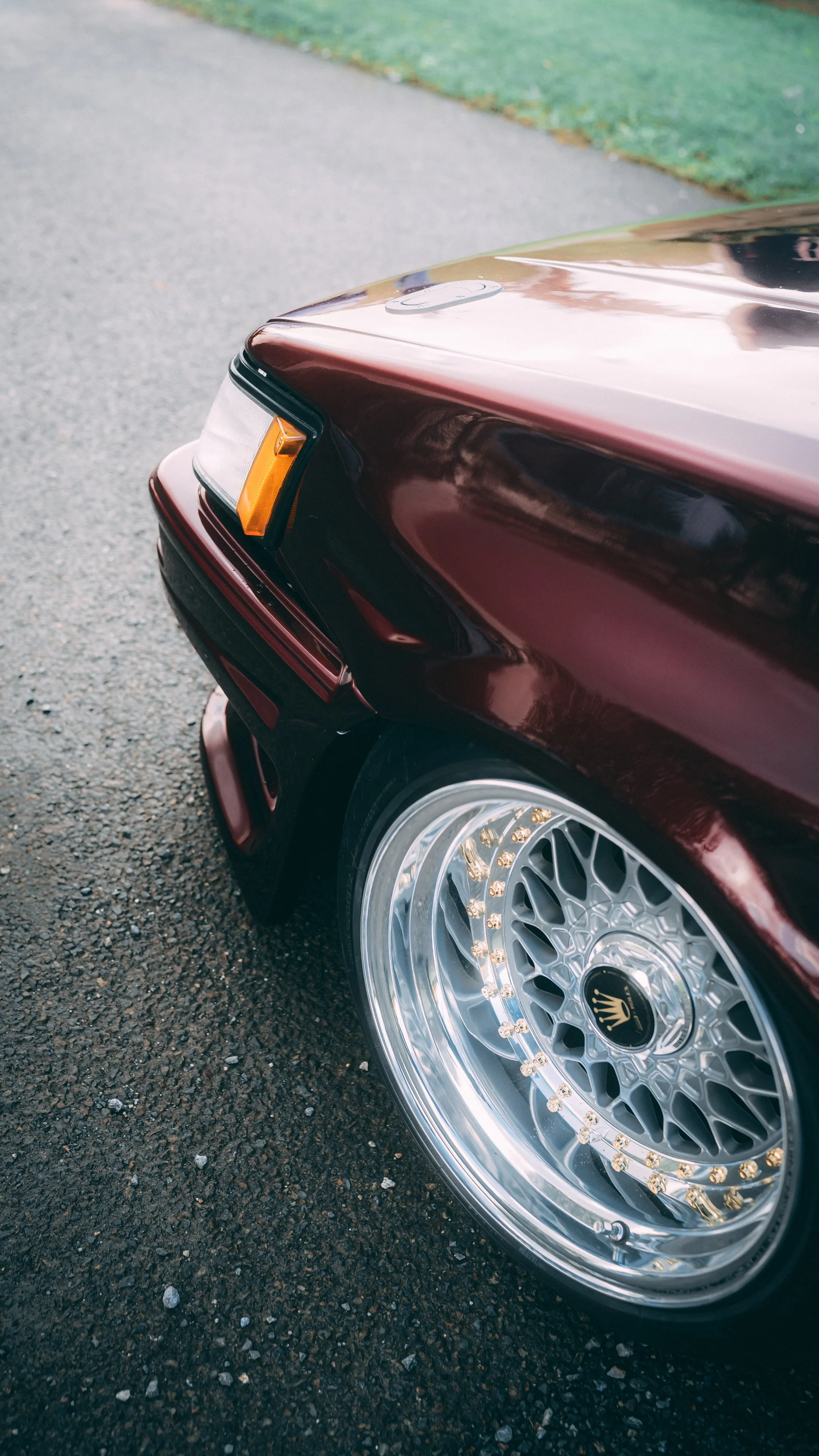 Close-up of a dark red car with a custom silver rim wheel on a pavement with green grass in the background.