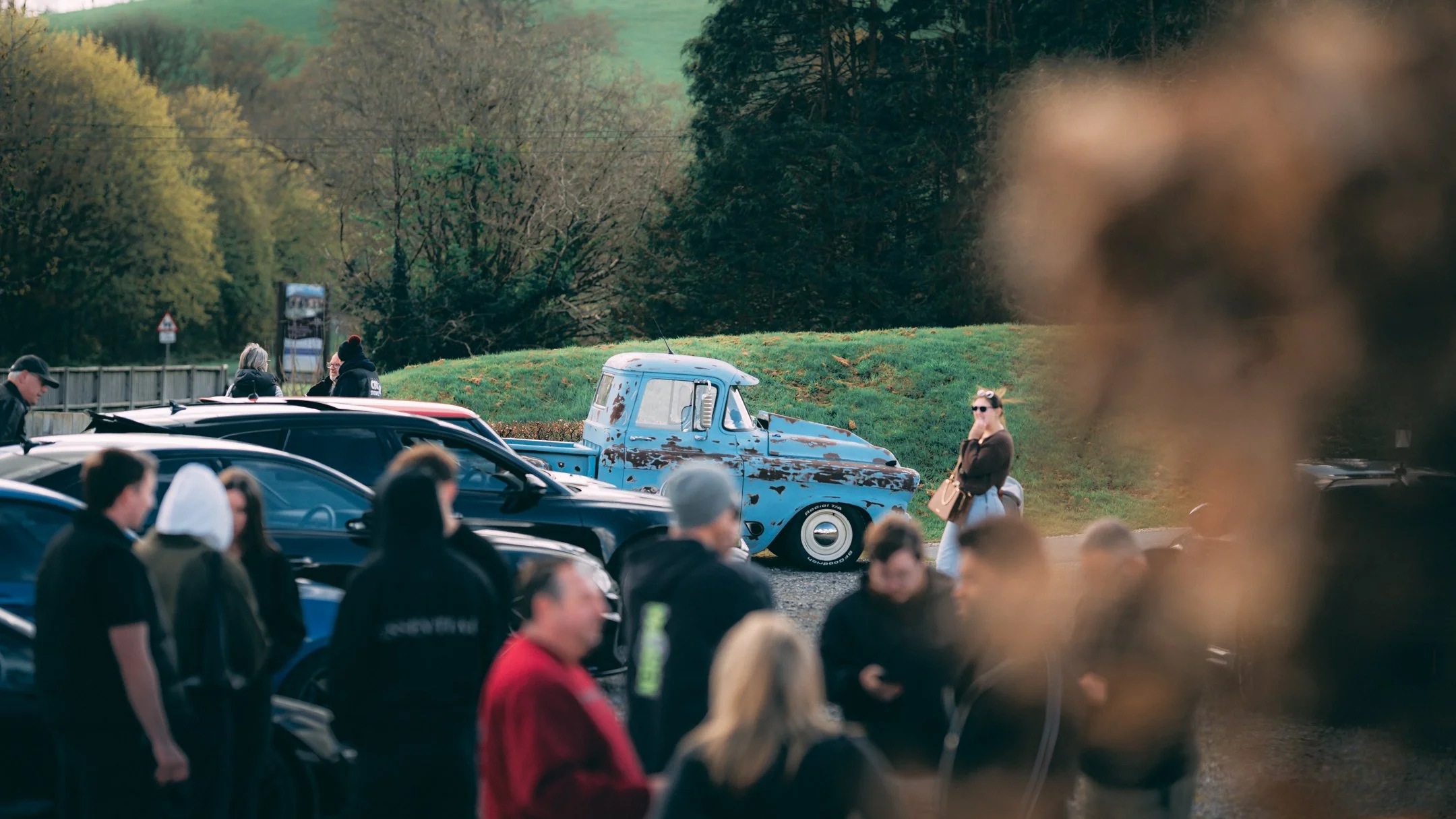 A crowd of people gathered outdoors near parked cars, including a weathered blue pickup truck, with trees and a grassy hill in the background.